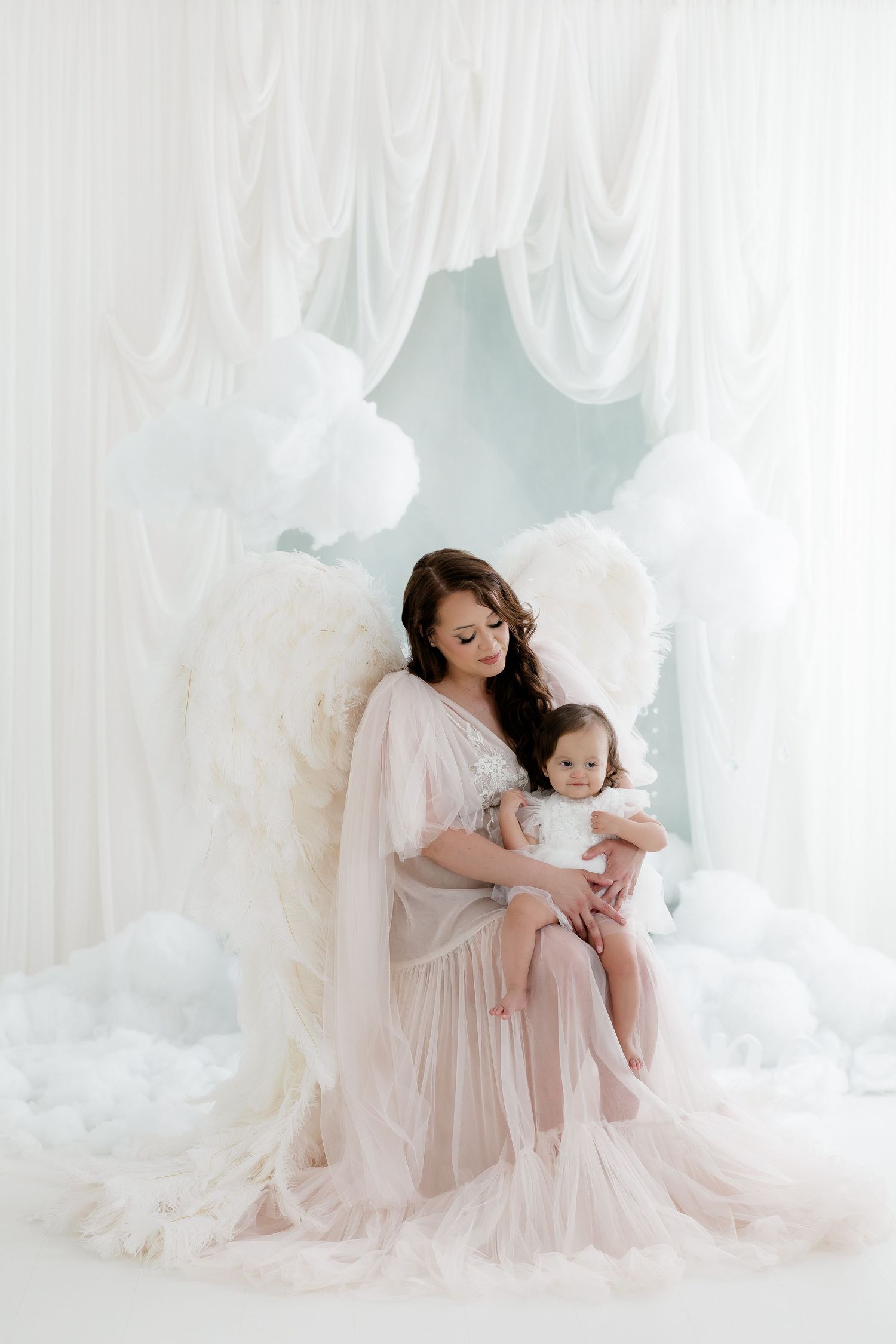 Mother wearing angel wings holds her baby girl amid white clouds and draping curtains in studio.