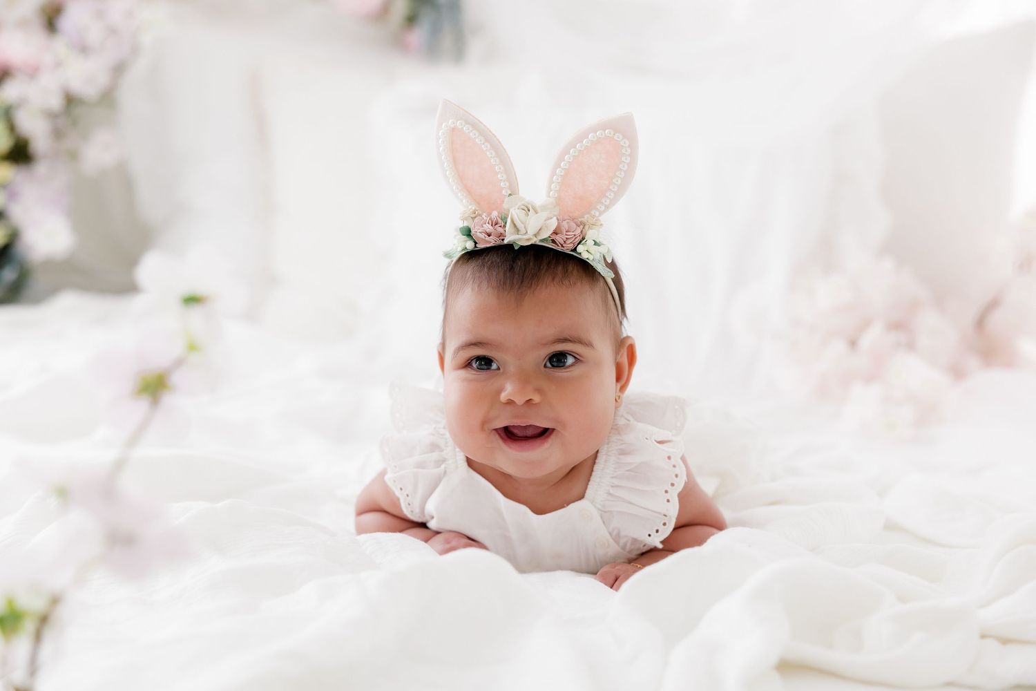 A baby wearing bunny ears and a white dress lies on soft white fabric surrounded by delicate floral decorations.