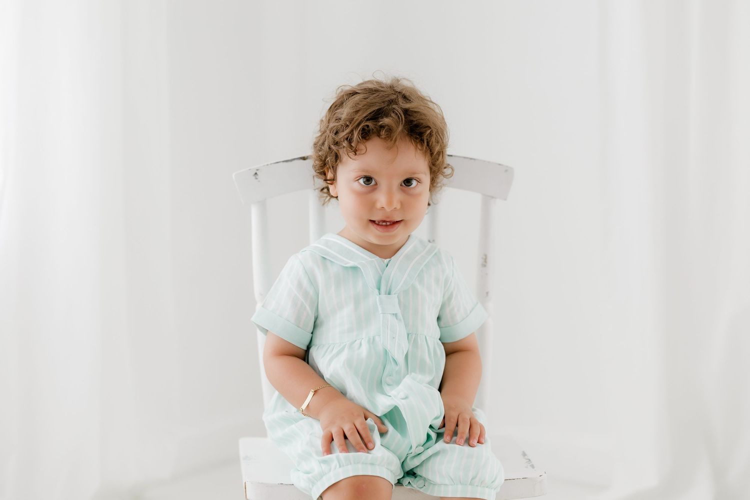 Baby in mint green outfit sits in white high chair with curly hair and happy expression.