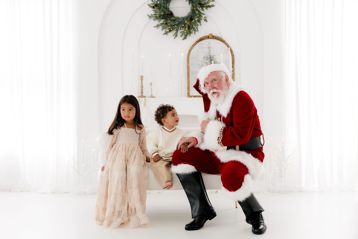 Two children pose with Santa Claus in a bright white holiday studio setting with a festive wreath backdrop.
