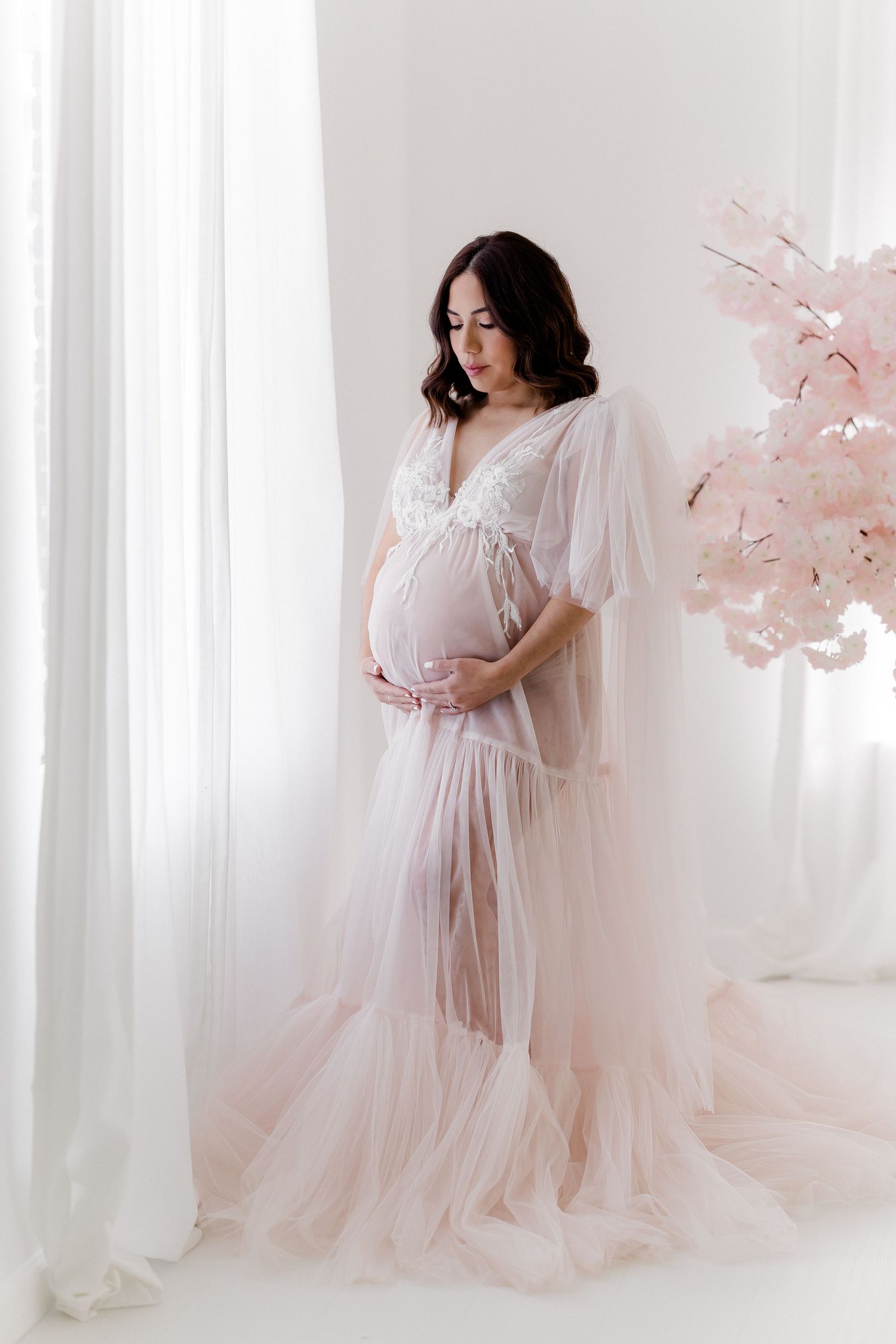 Ethereal maternity photoshoot featuring flowing pink tulle gown against white curtained backdrop.