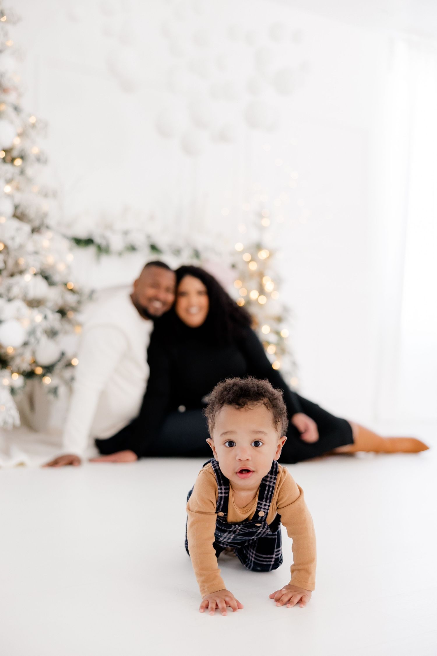 Baby crawling on white floor during holiday photoshoot with decorated Christmas trees in background.