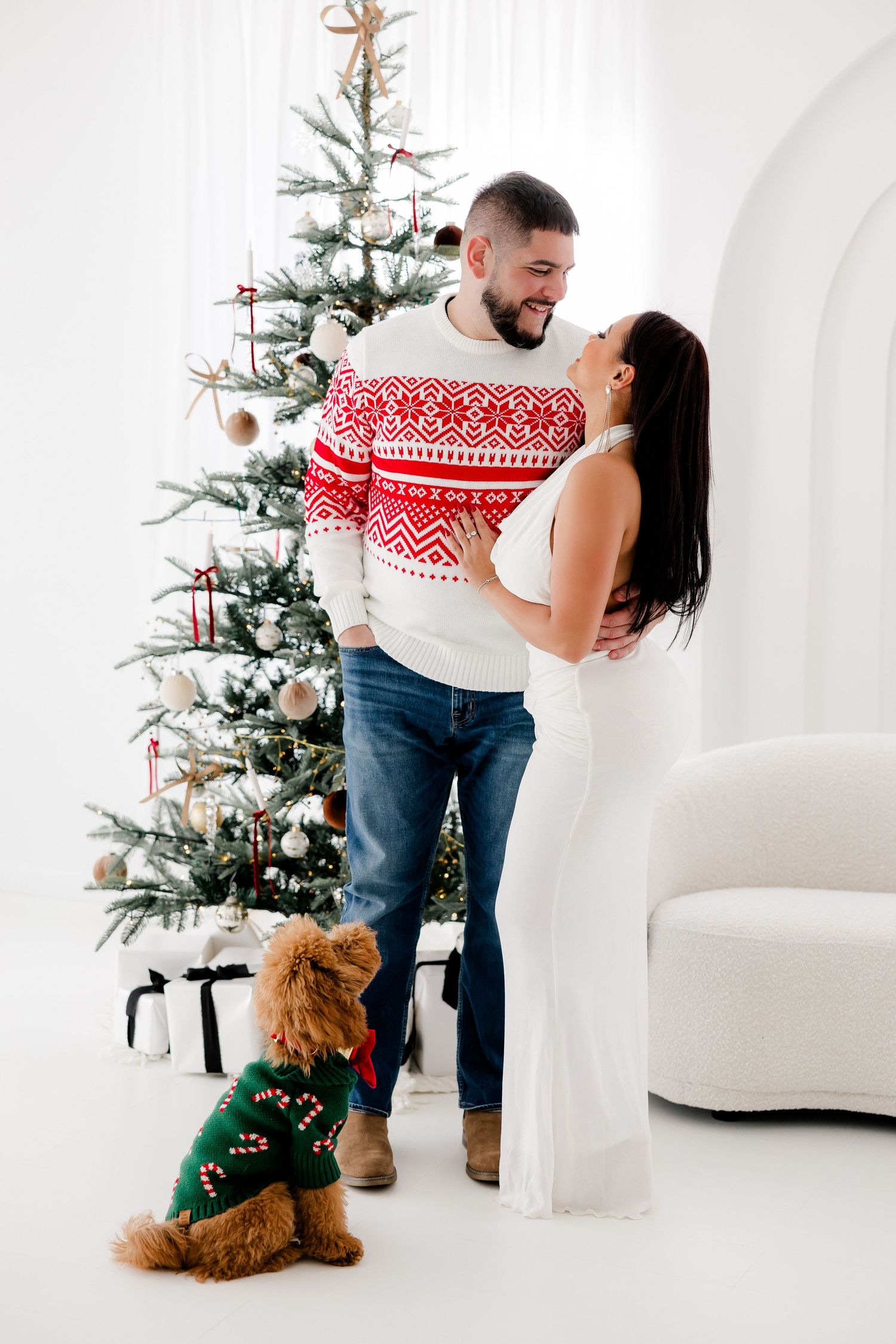 Couple in festive winter outfits sharing playful moments by Christmas tree with their dog.nj holiday mini sessions