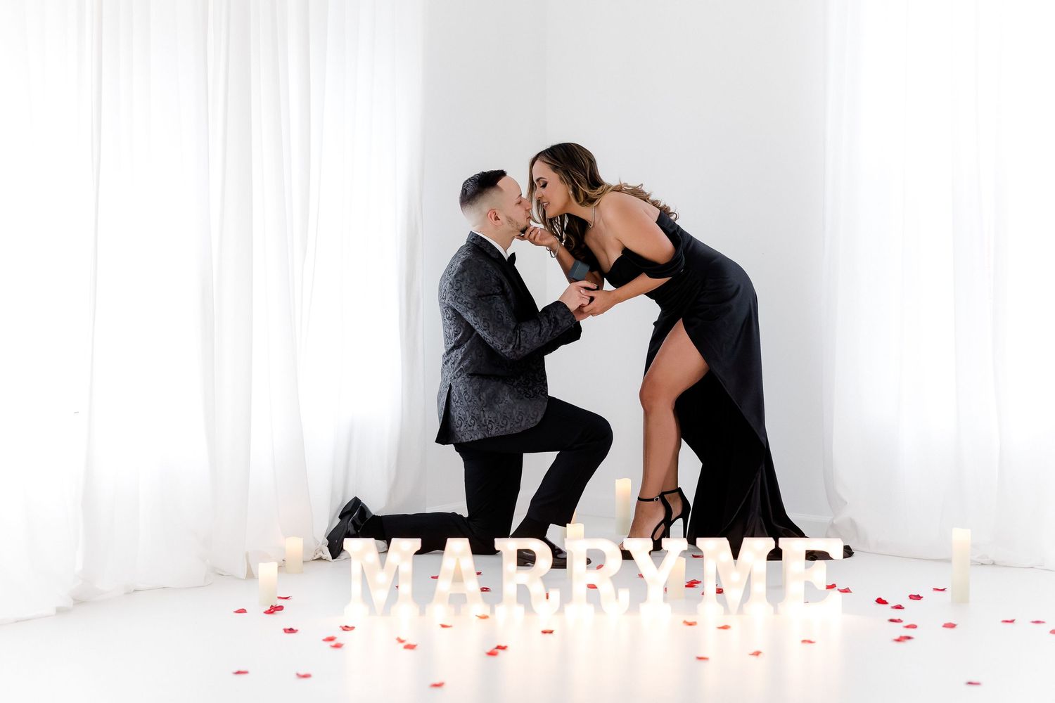A romantic proposal scene with candles and rose petals scattered on white floor as couple shares intimate moment.