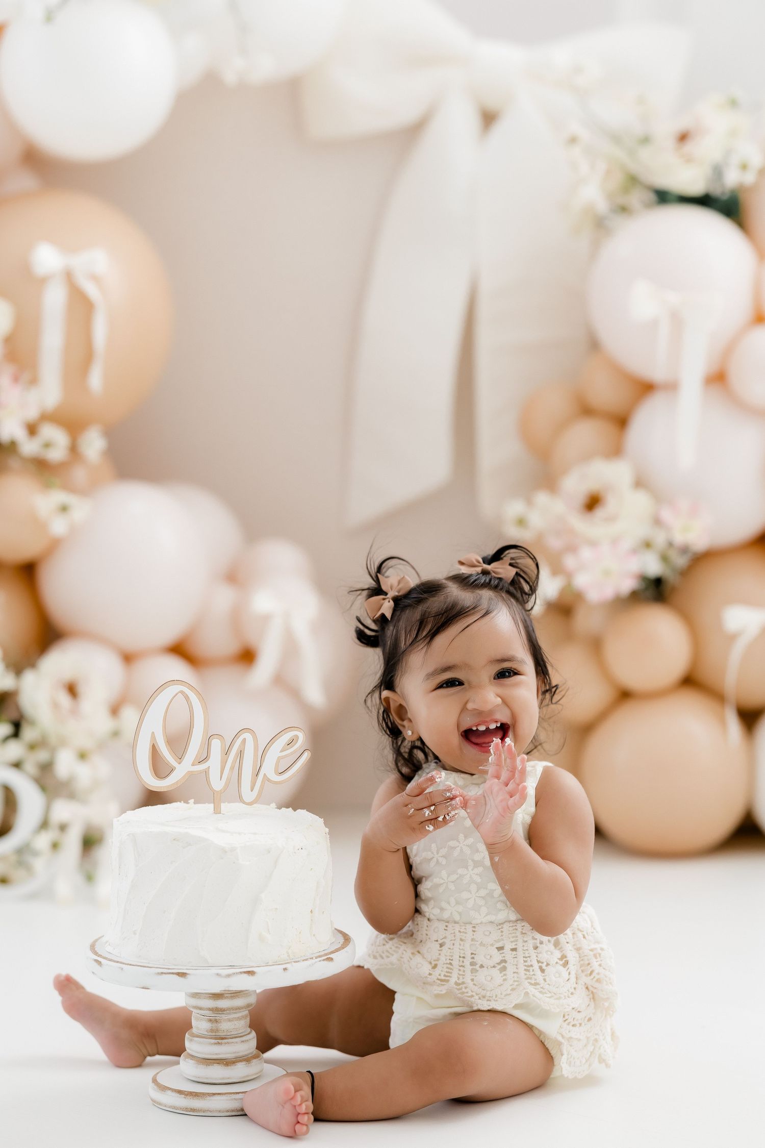 A baby in a white lace dress sits and laughs during a first birthday cake smash photoshoot with neutral balloon decor.