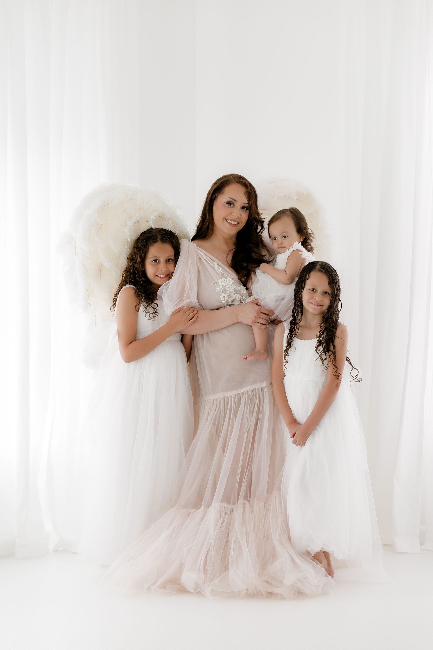 A pregnant woman in a white gown poses with three daughters in white dresses and angel wings in a bright studio.