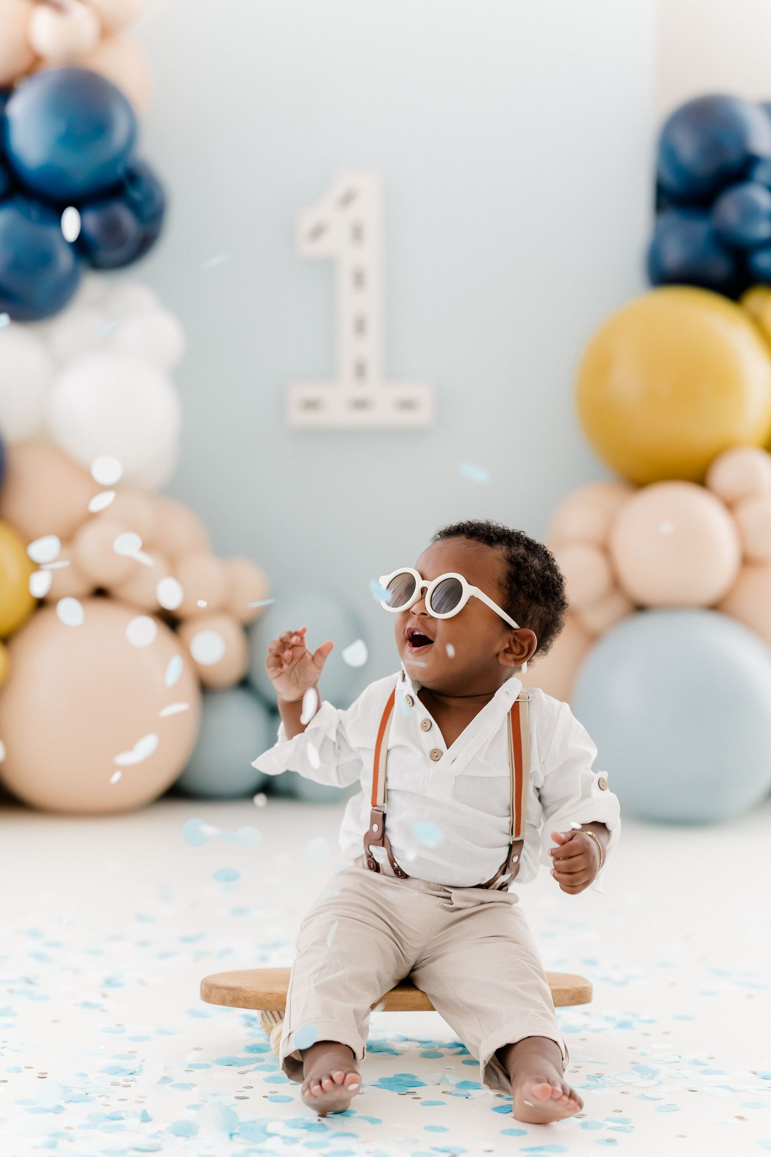 Cake smash photo session with blue and gold balloons and confetti on white floor.