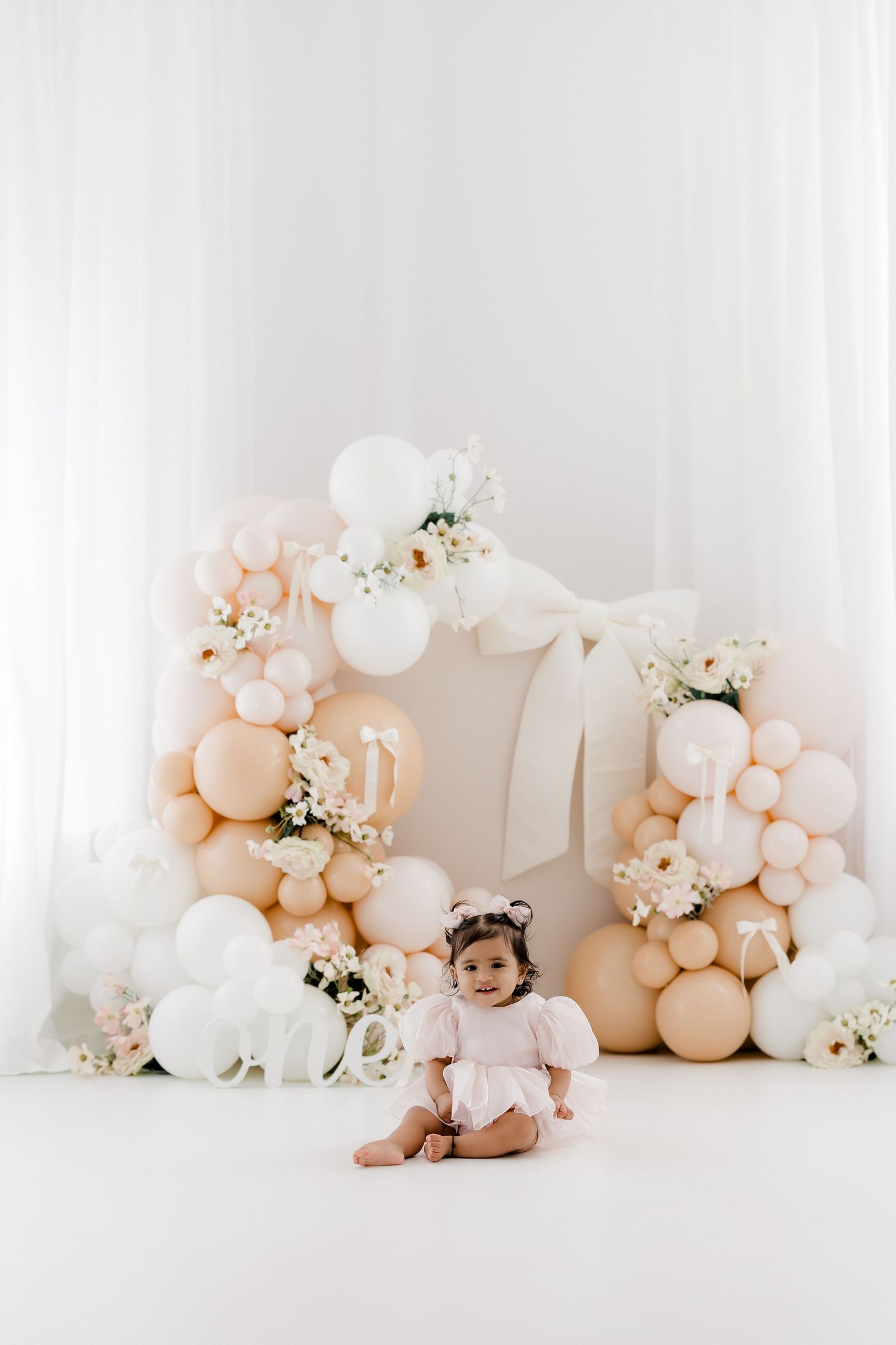 Elegant baby photo setup with blush and white balloon garland, floral accents, and white backdrop in a minimalist studio setting.