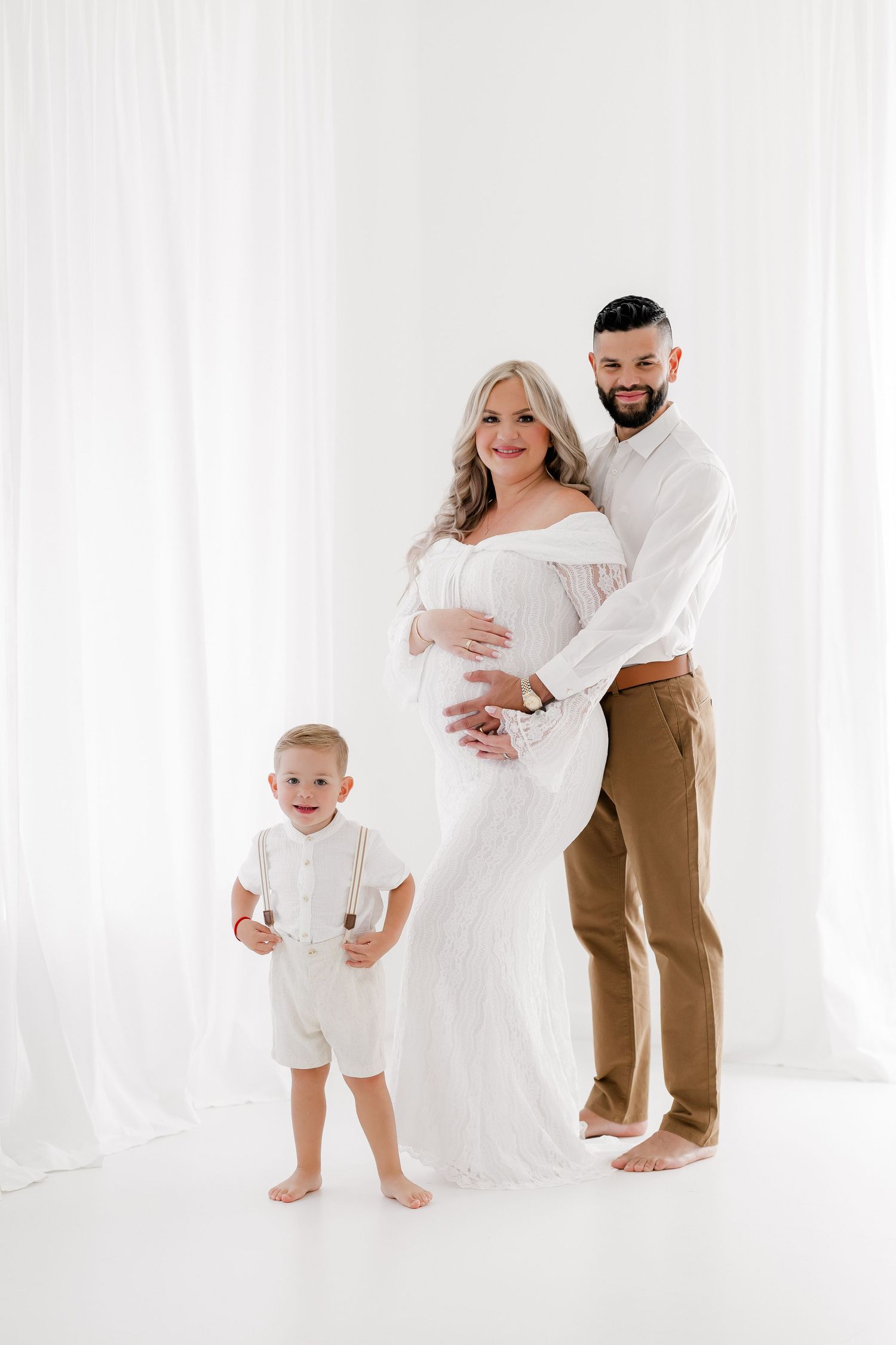 Family maternity photo session in an all-white studio with everyone wearing white and tan clothing.