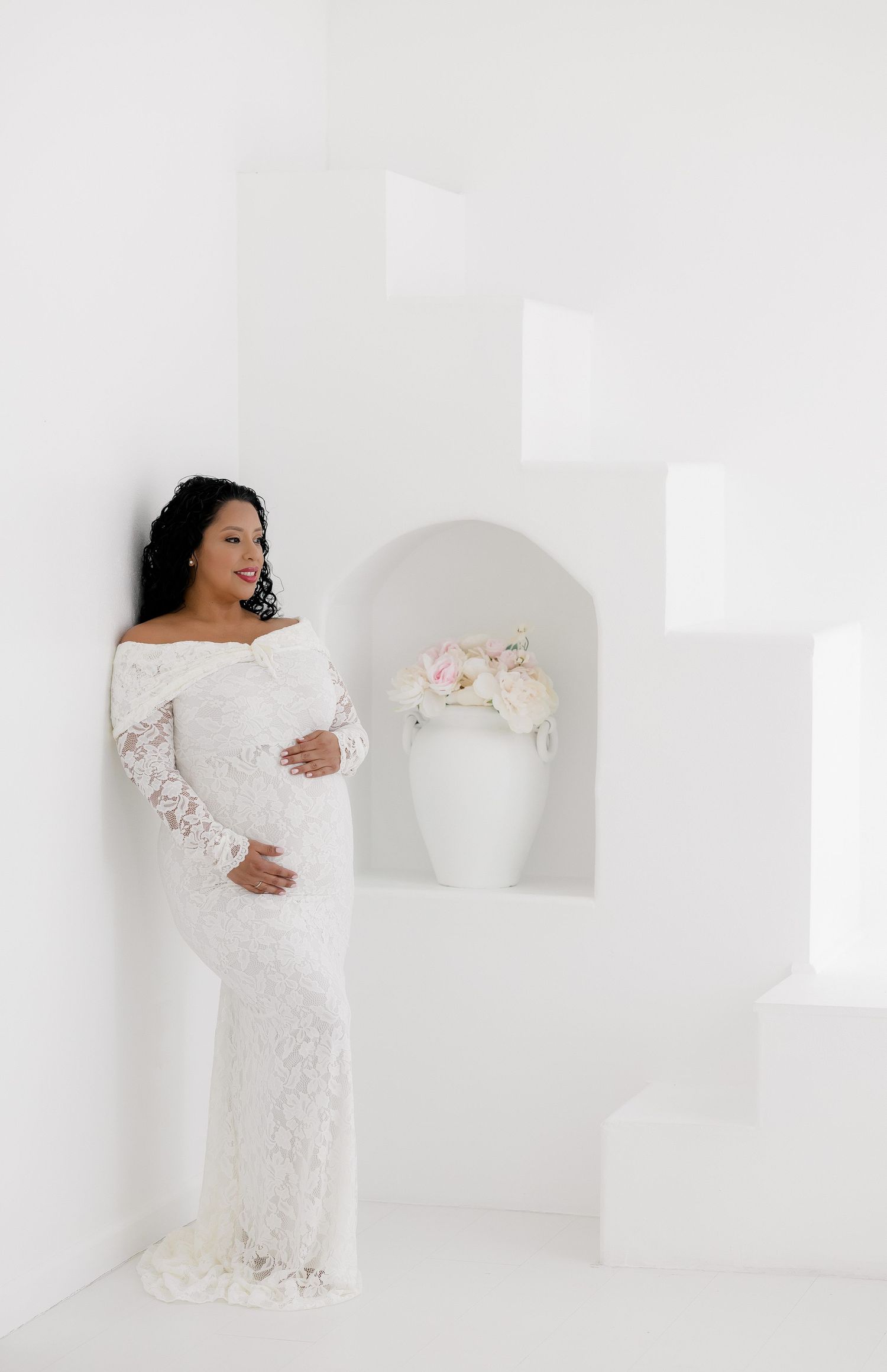 A person in a white lace dress poses gracefully against a minimalist white backdrop in a bridal photoshoot.