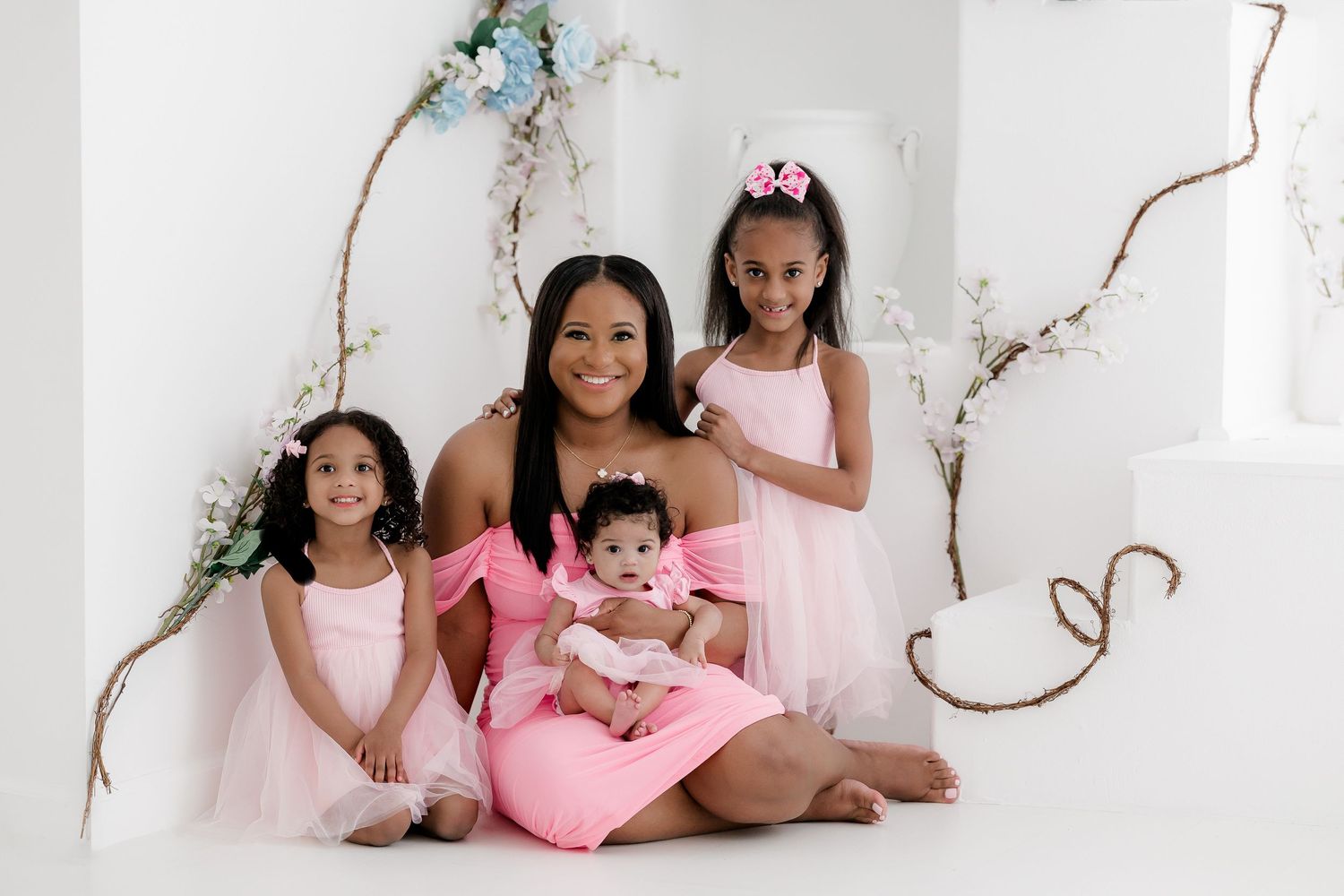 A group in soft pink dresses poses together against a white backdrop with delicate floral branches.