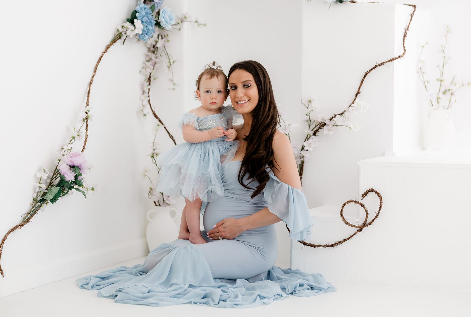 Two people in matching light blue dresses pose together against a white backdrop with decorative twigs and flowers.