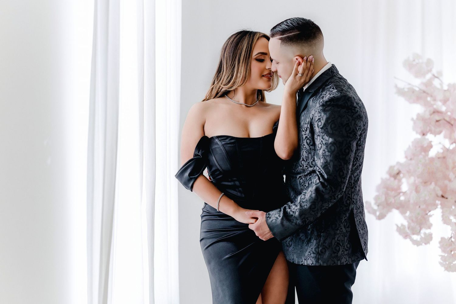 A romantic couple in elegant black attire share intimate moments in a bright white room with natural lighting.