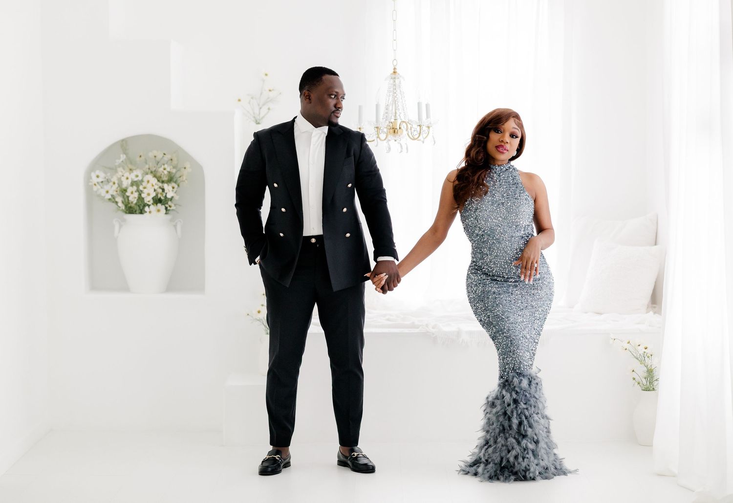 A couple in formal attire poses together in an elegant white studio setting with floral decor.