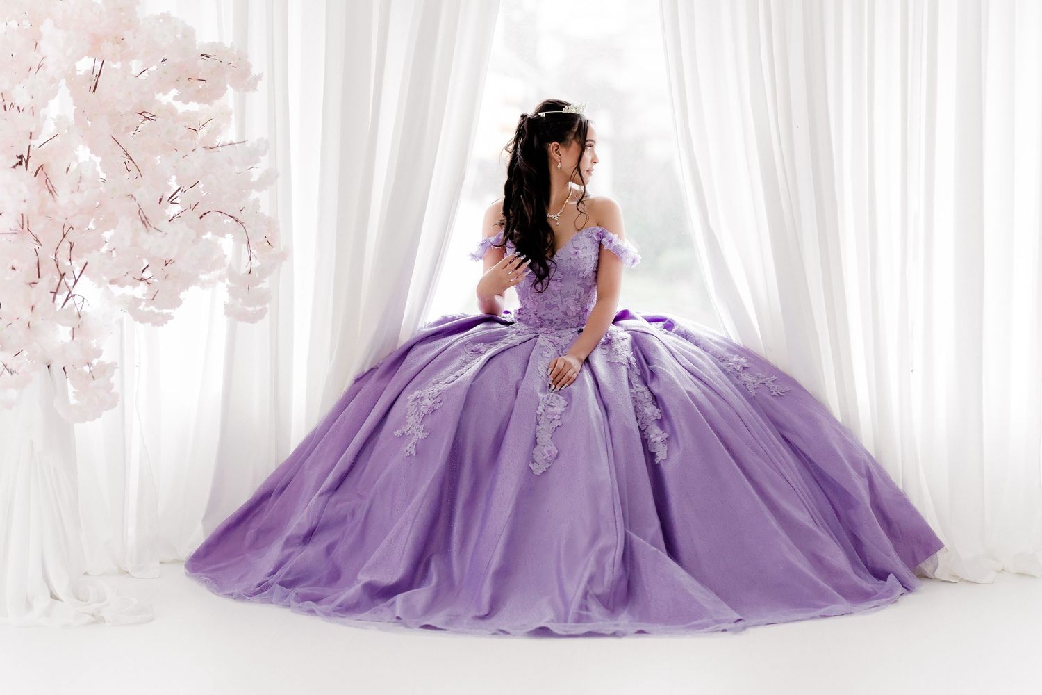 A model in a flowing purple ball gown poses gracefully against a white backdrop with romantic lighting.