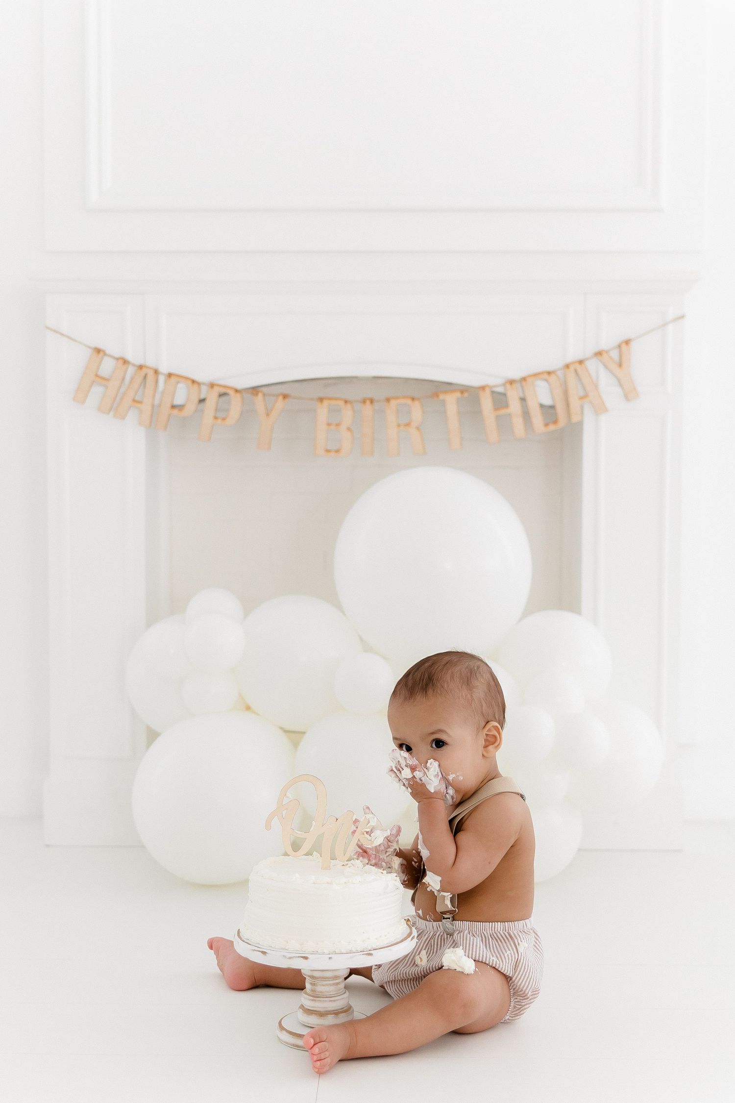 Happy birthday cake smash photo session with white balloons and festive banner.