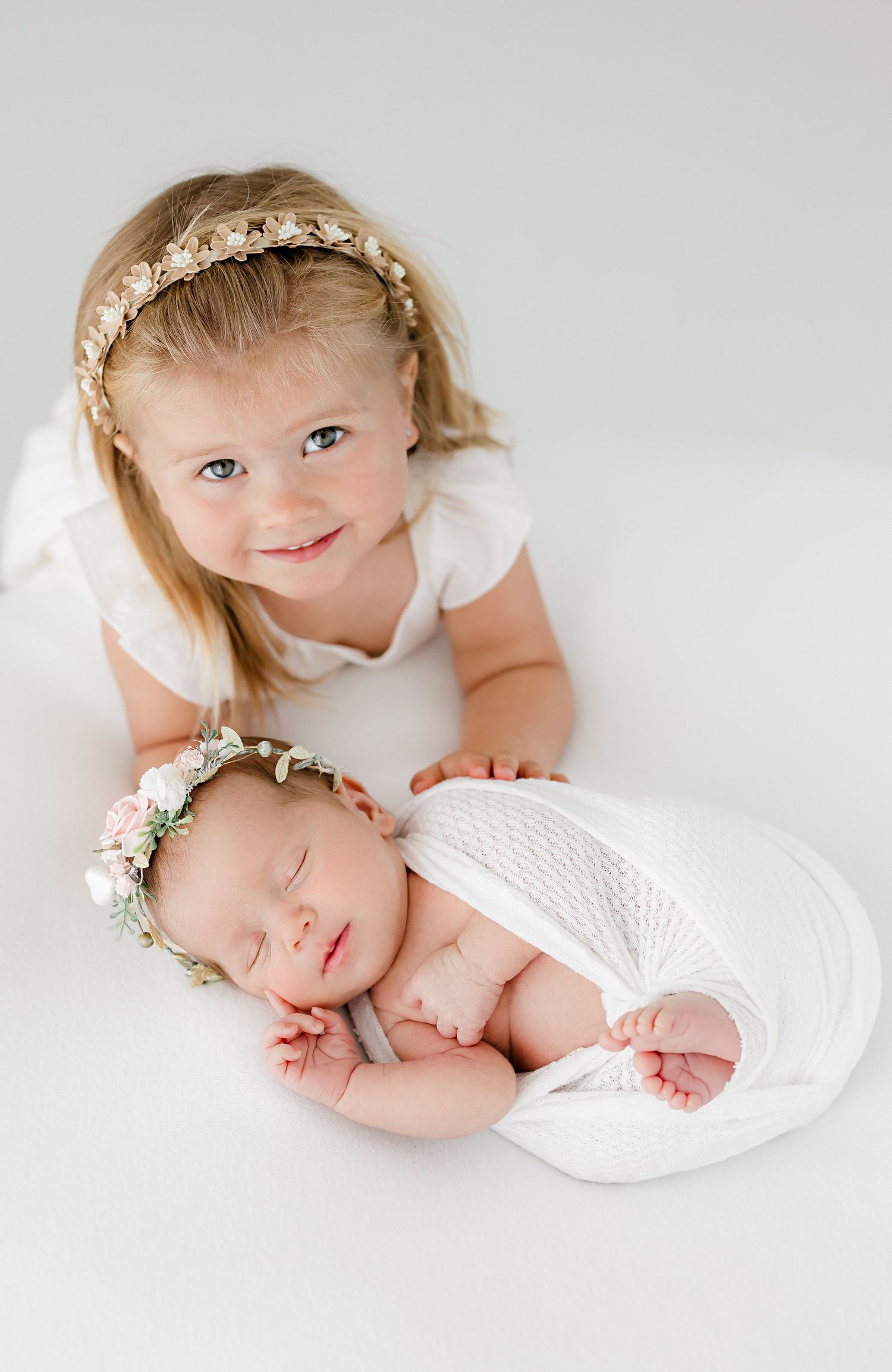Sweet newborn photography session showing siblings in soft white clothing and delicate floral headpieces.