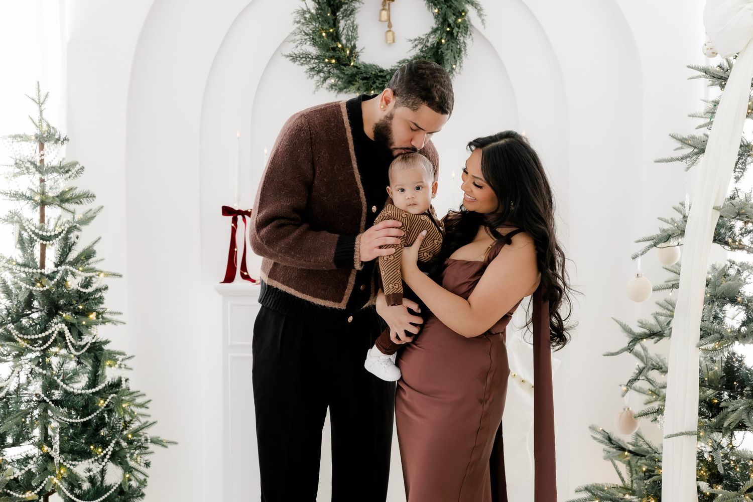 Family Christmas photo with parents holding baby near decorated trees and white arch backdrop.