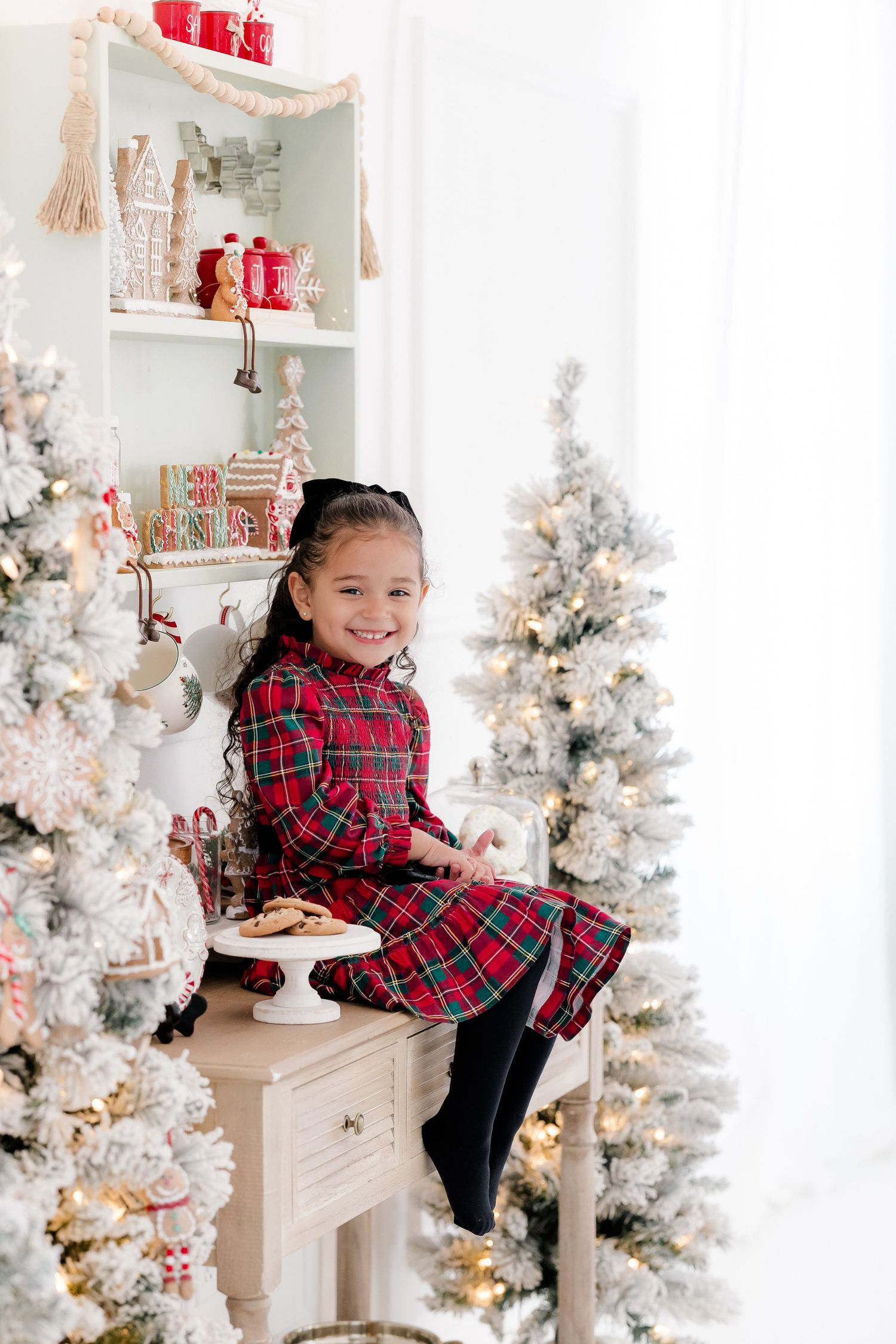Young girl in red plaid dress sits smiling among flocked Christmas trees with holiday decorations and cookies nearby.