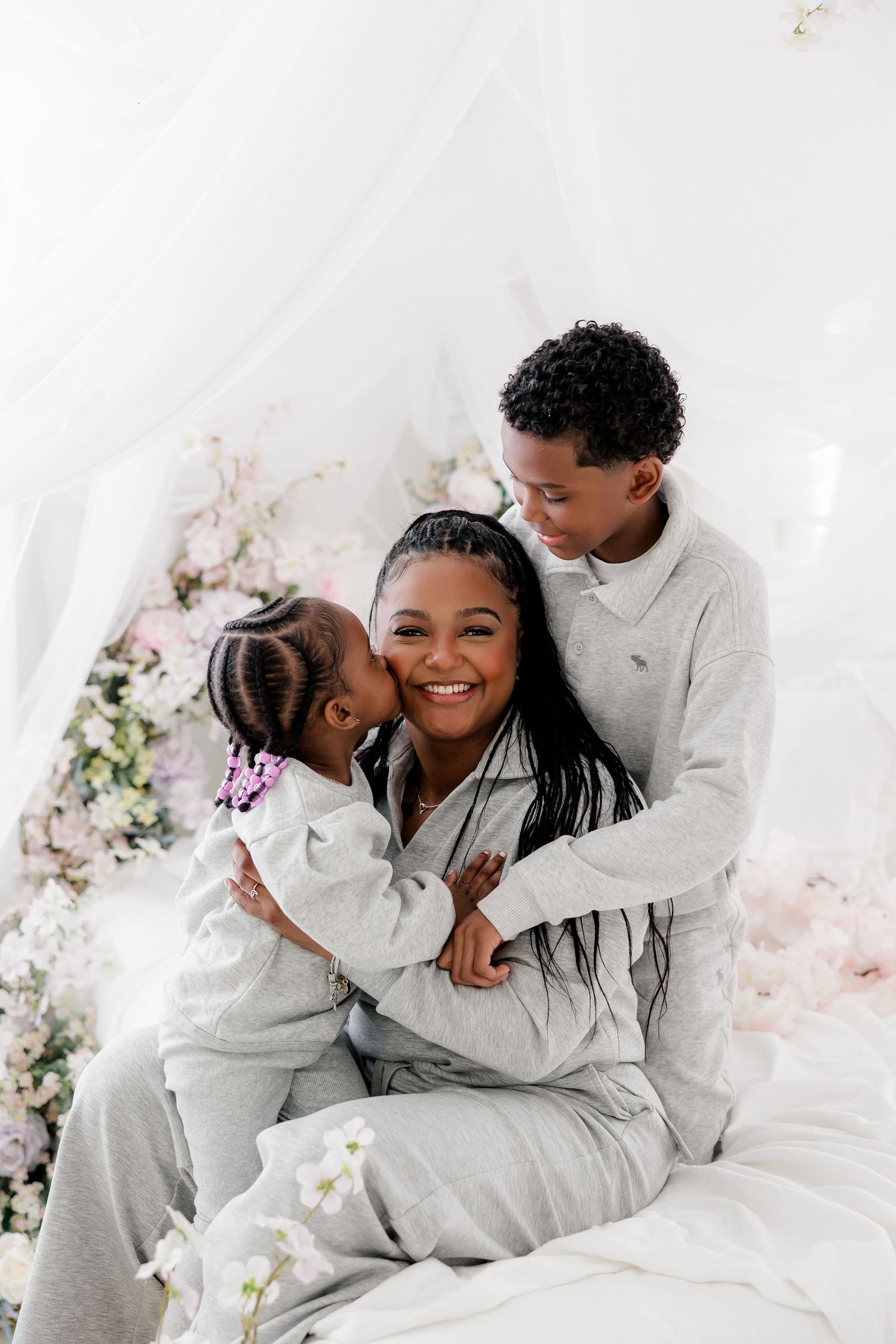 A heartwarming family portrait in matching grey outfits sharing loving embraces against a white floral backdrop.
