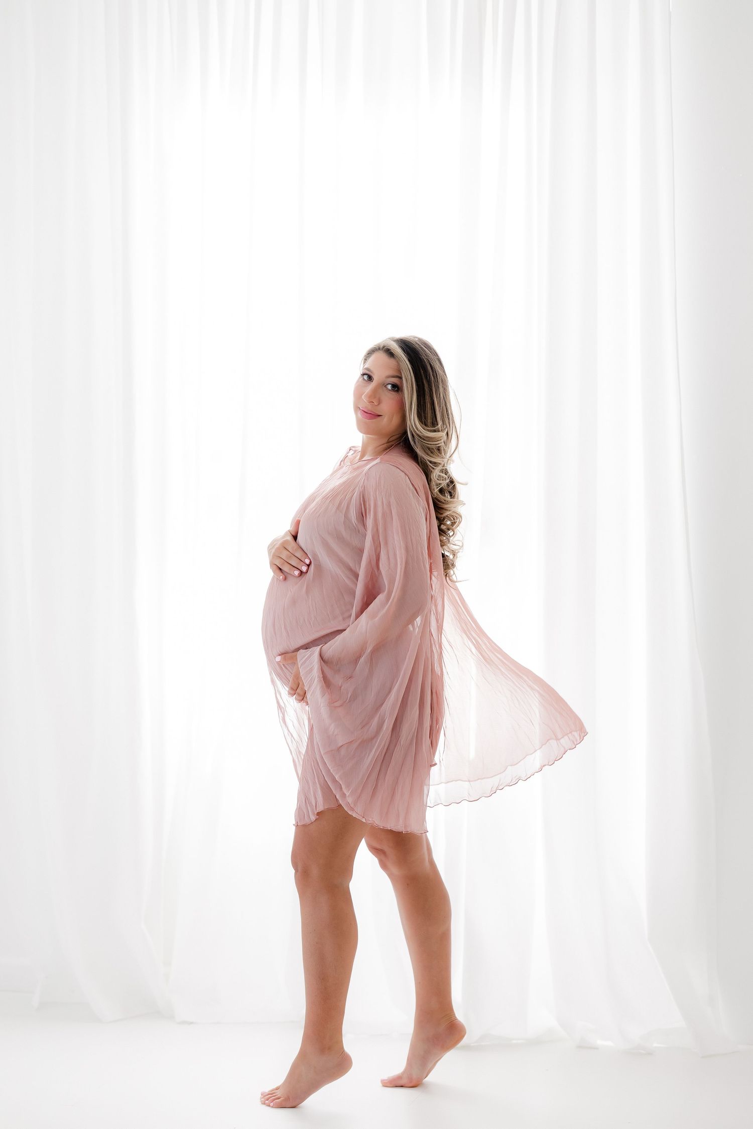 Four boudoir poses in a white studio setting wearing a sheer pink robe against white curtains and natural lighting.