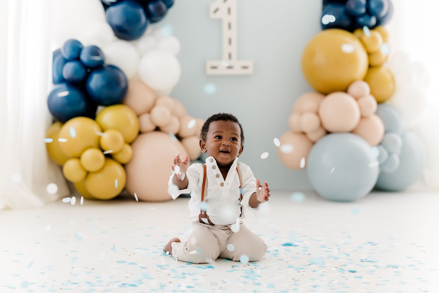 First birthday setup with blue and gold balloon arch and scattered confetti.