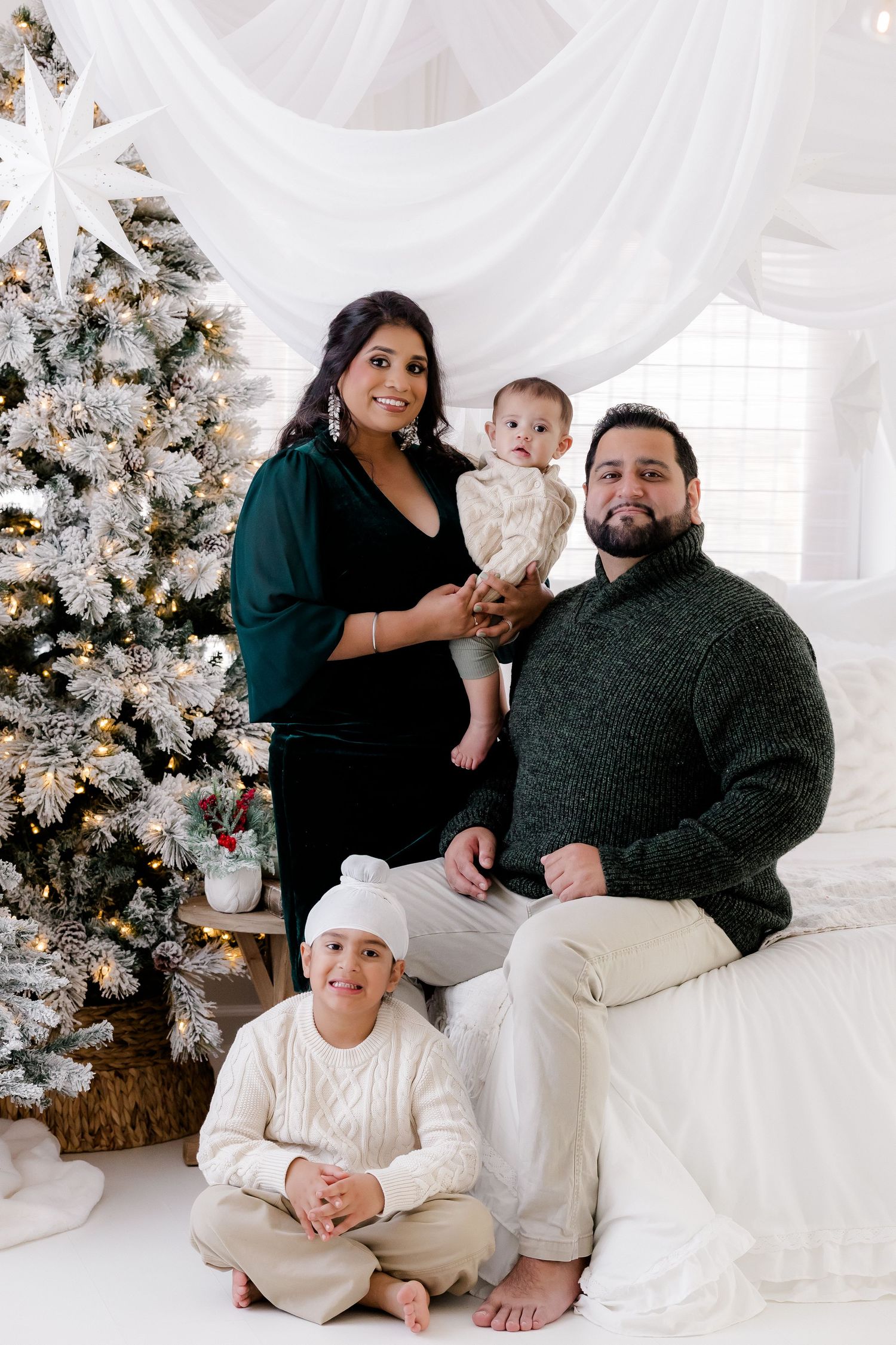 Family of four posing for a Christmas portrait near a flocked tree in a bright white studio setting.