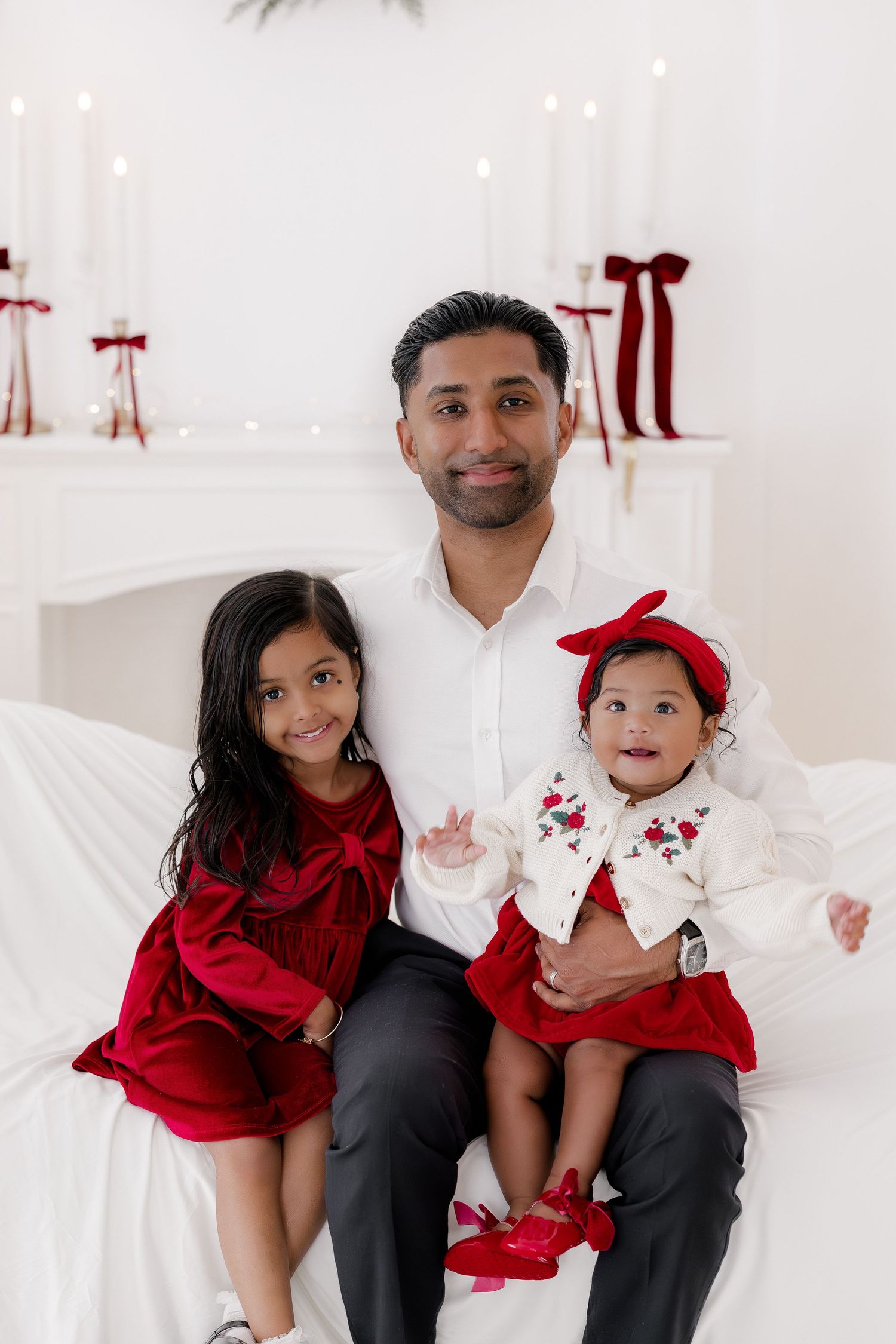 A father poses with two young daughters in red holiday outfits against a white backdrop with candles and festive decor.