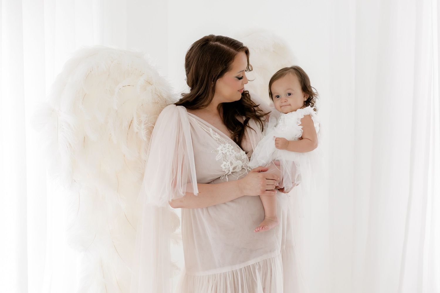 Pregnant woman with angel wings holding a baby girl in white dresses in a bright studio setting.