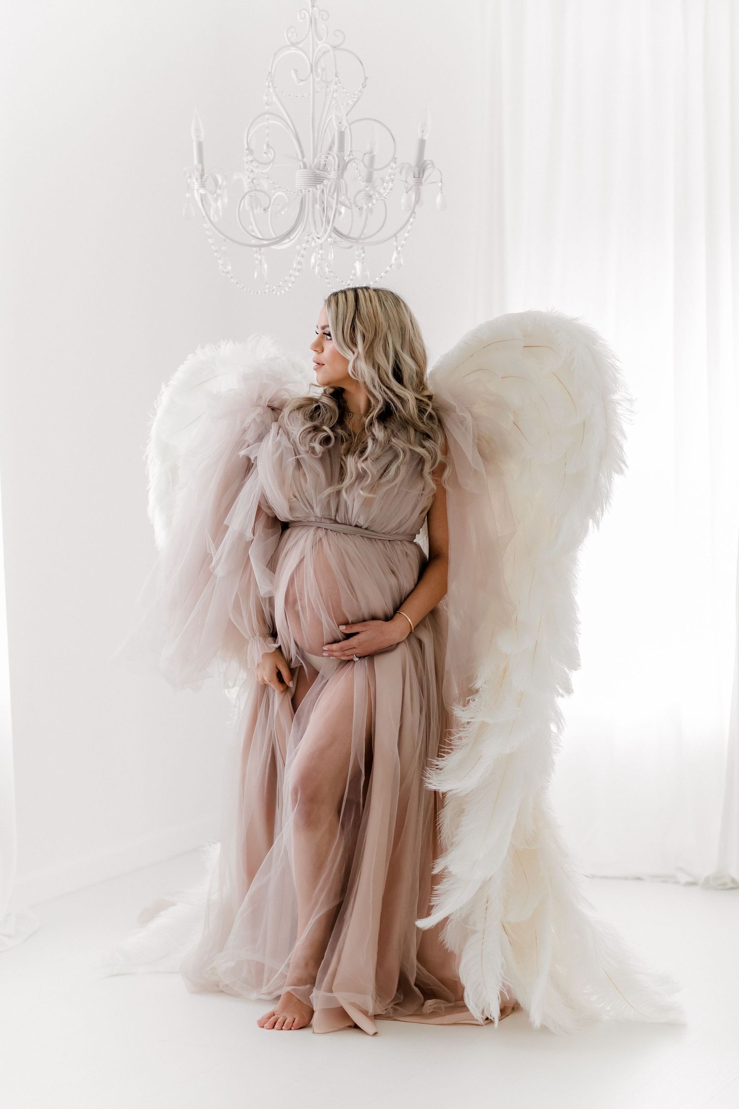 Ethereal maternity portrait with angel wings and blush gown under crystal chandelier.