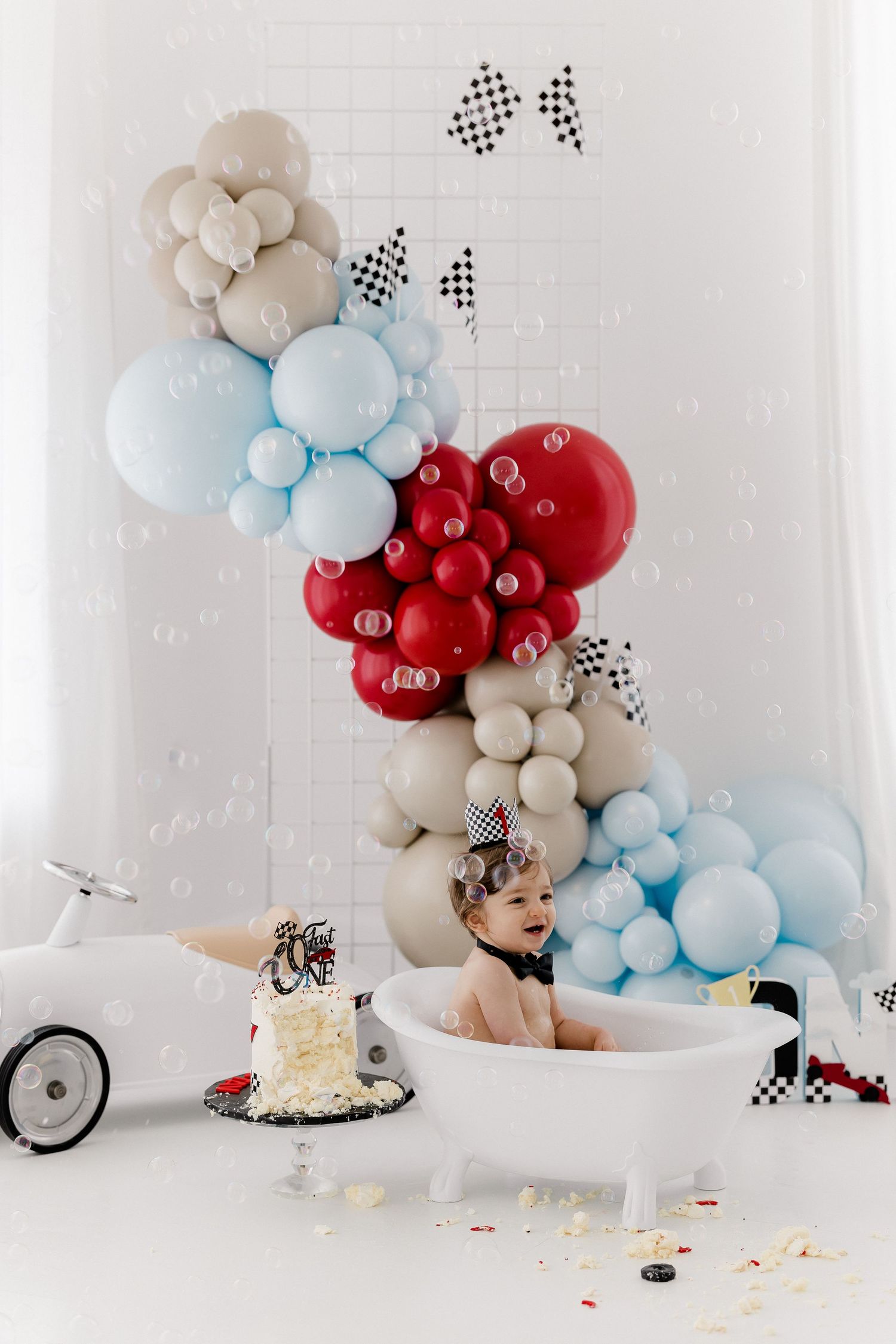 Baby's cake smash photoshoot with red and blue balloon garland and white brick backdrop.