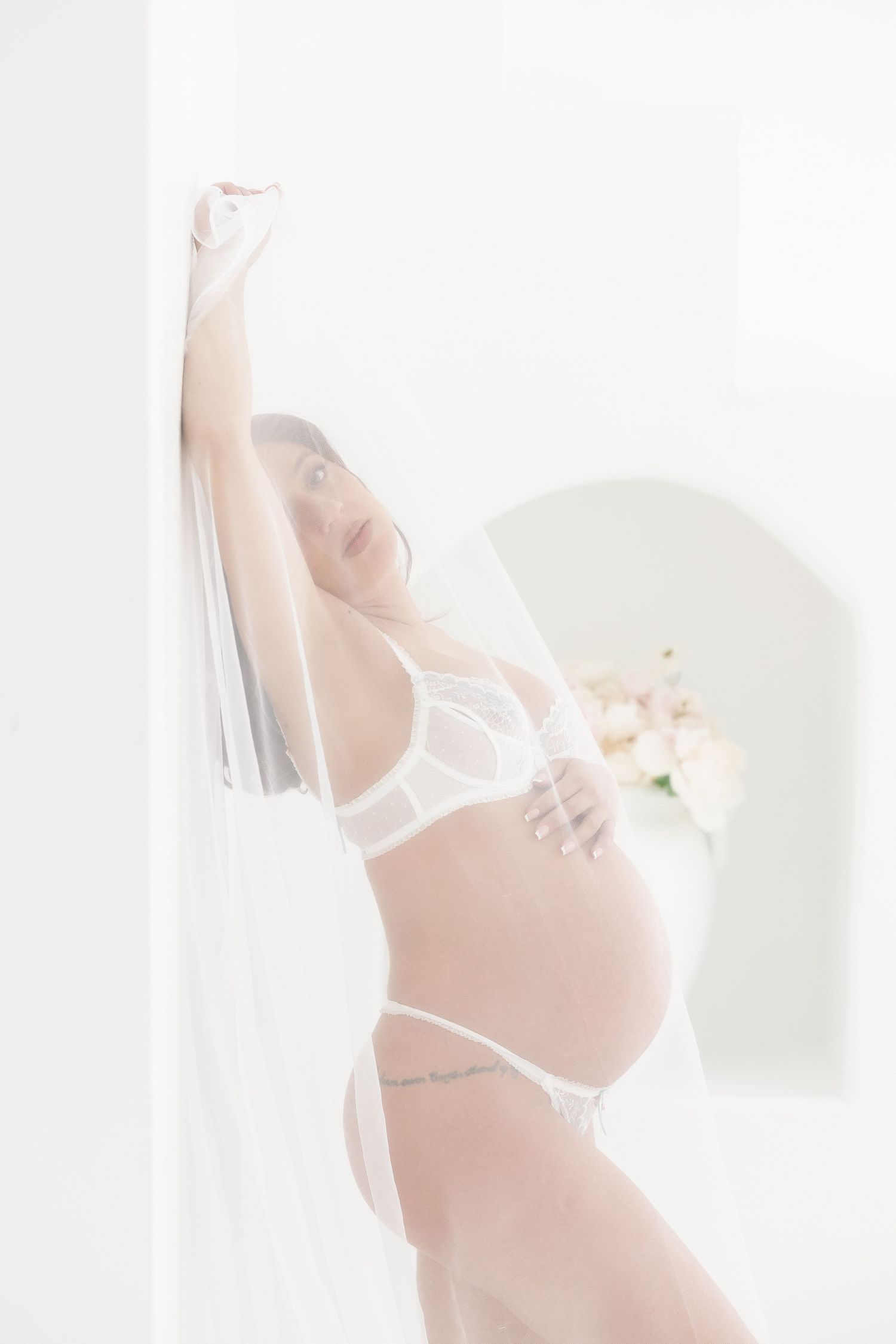 A pregnant woman in white lingerie poses gracefully against a light background during a maternity photoshoot.