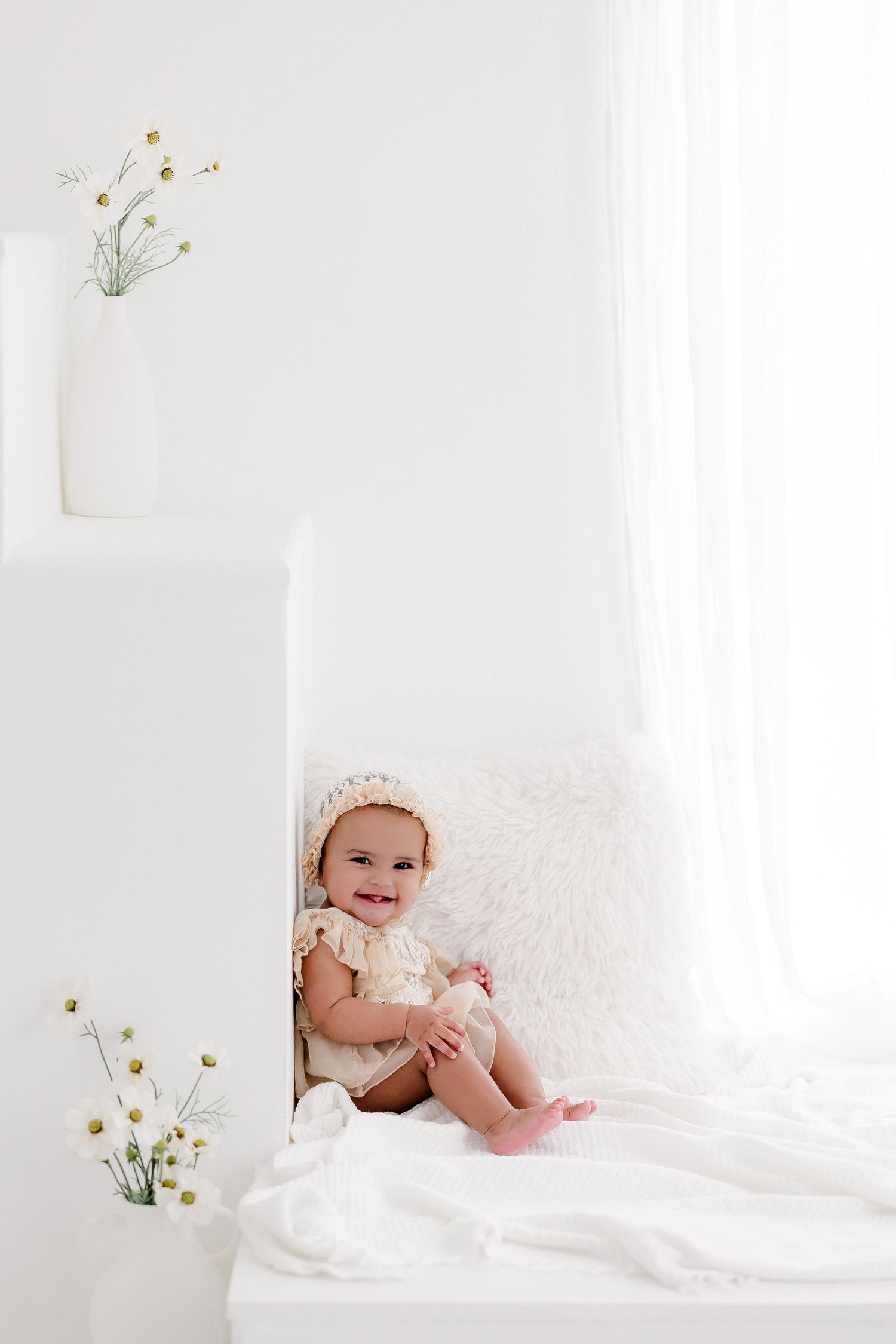 A baby wearing a white lace bonnet lies on white bedding while smiling and playing with delicate yellow flowers.
