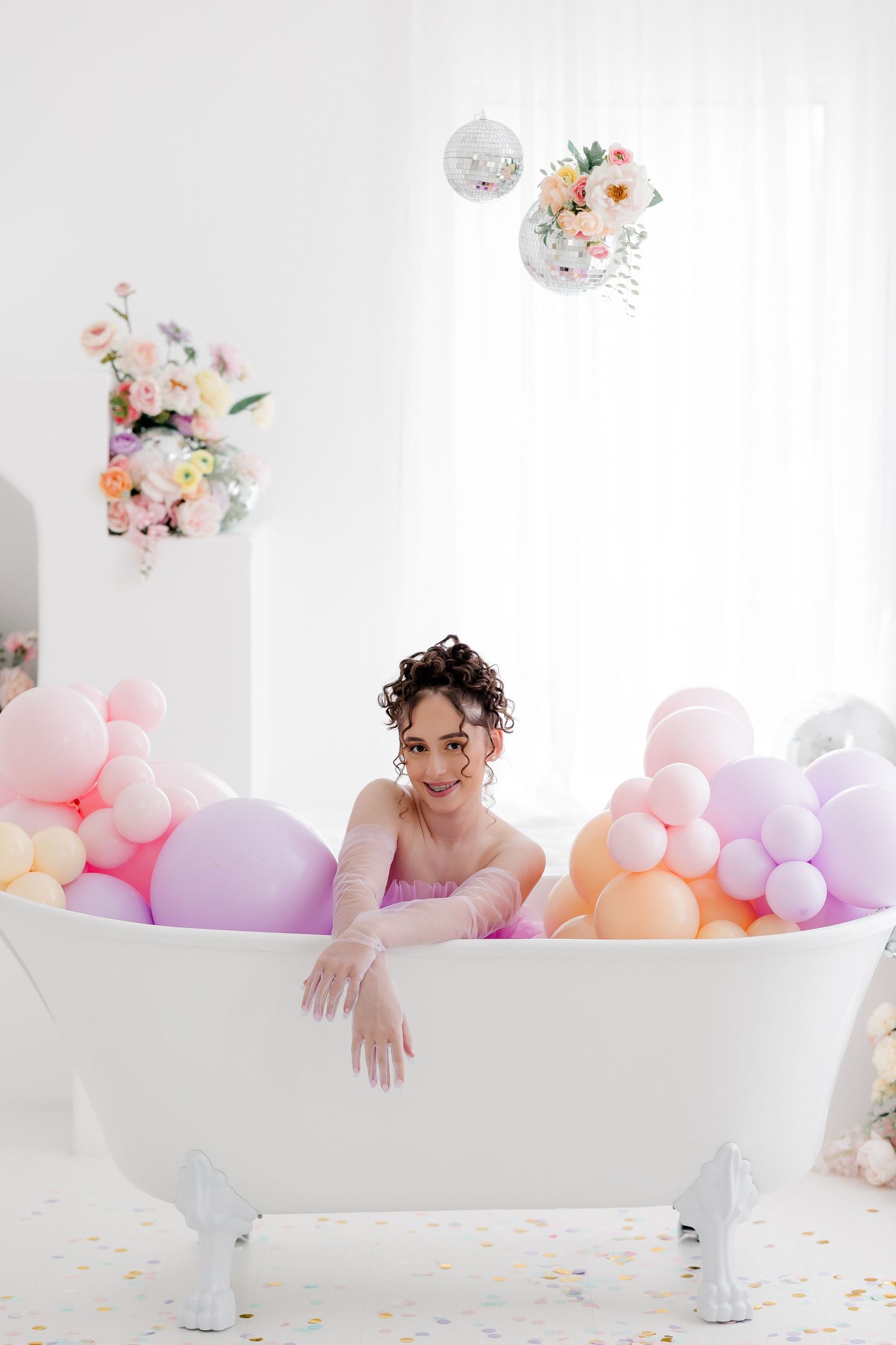 A bathtub filled with pastel balloons and floral decorations creates a whimsical and dreamy photography setup.