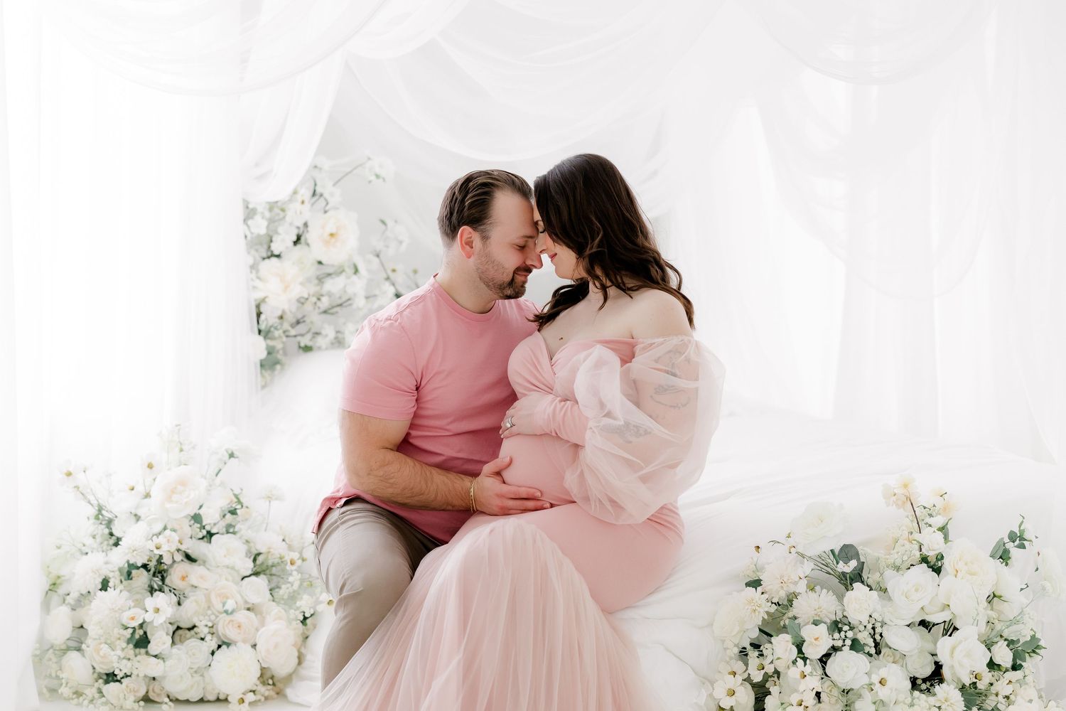 Romantic couple in pink and white outfits share intimate moments against floral backdrop during photo session.