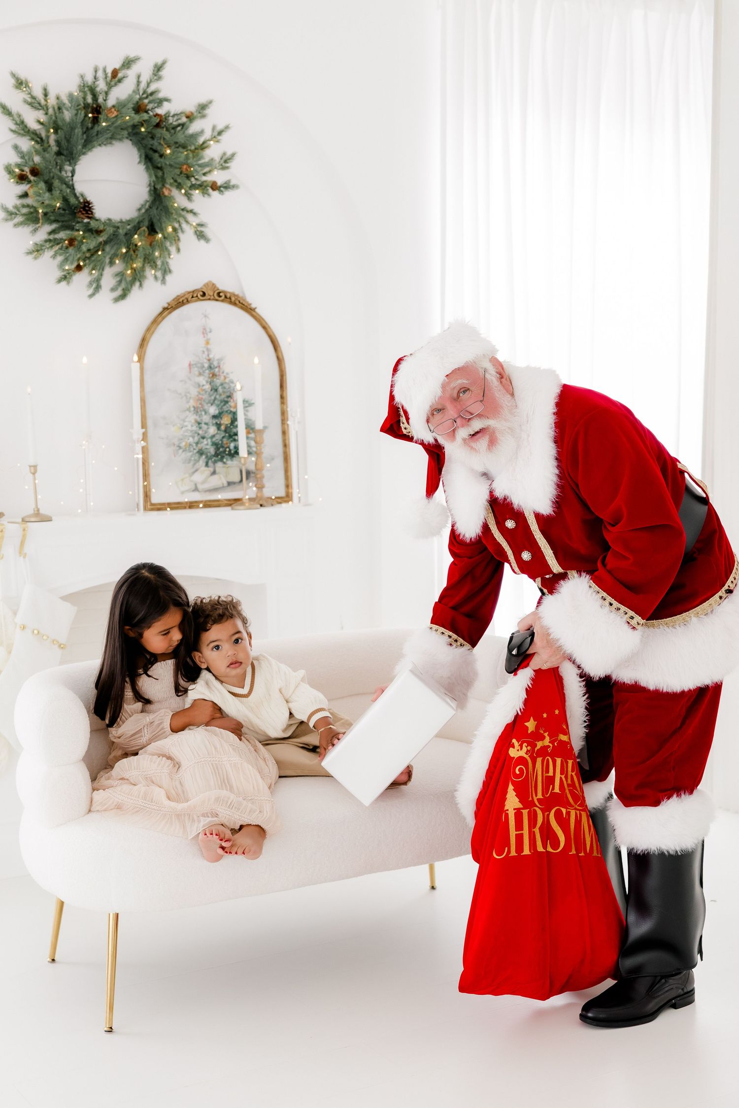 Santa Claus handing gifts to two children sitting on a white sofa in a festive decorated living room.