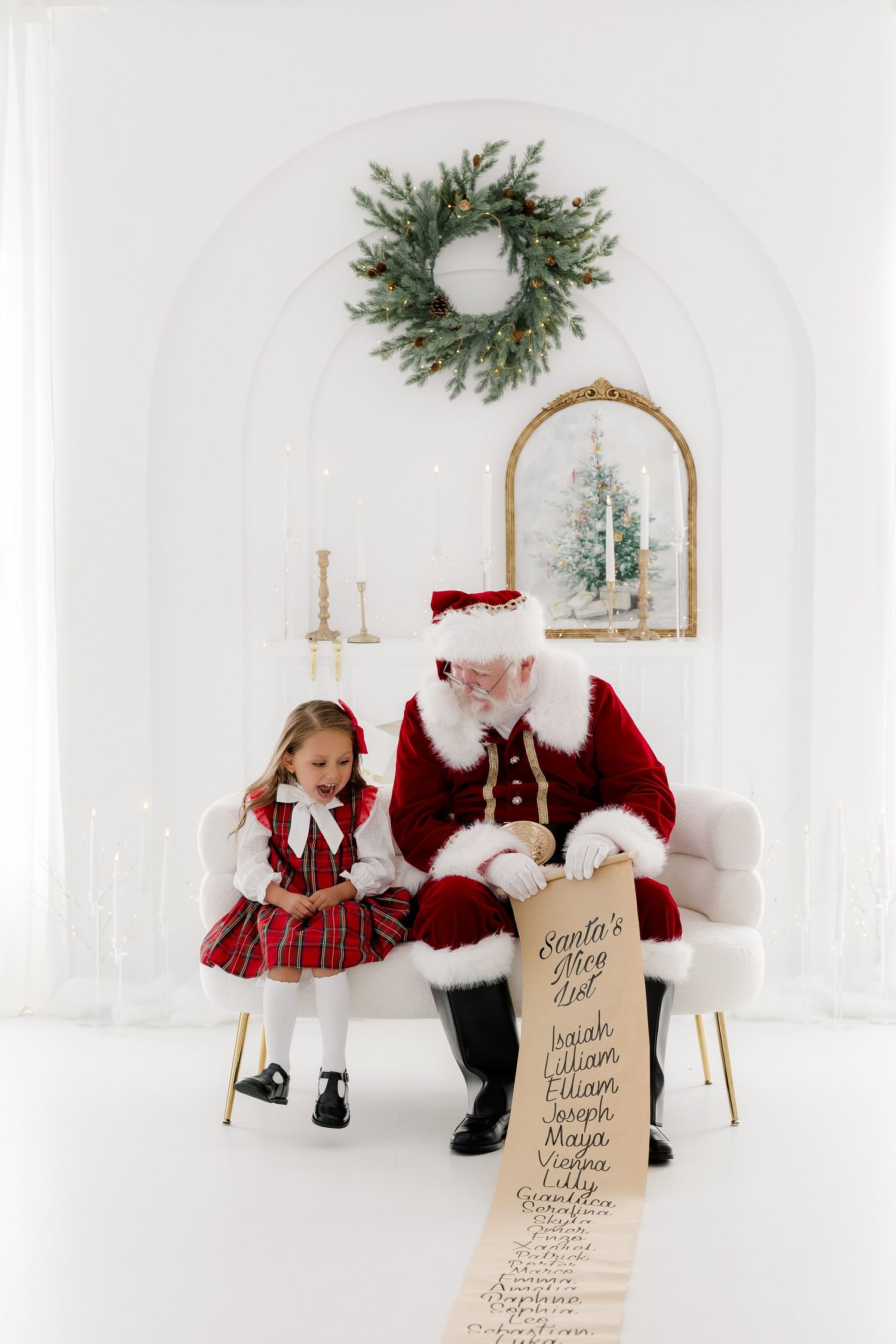 A little girl in plaid dress sits with Santa Claus reviewing his naughty and nice list in a white holiday setting.