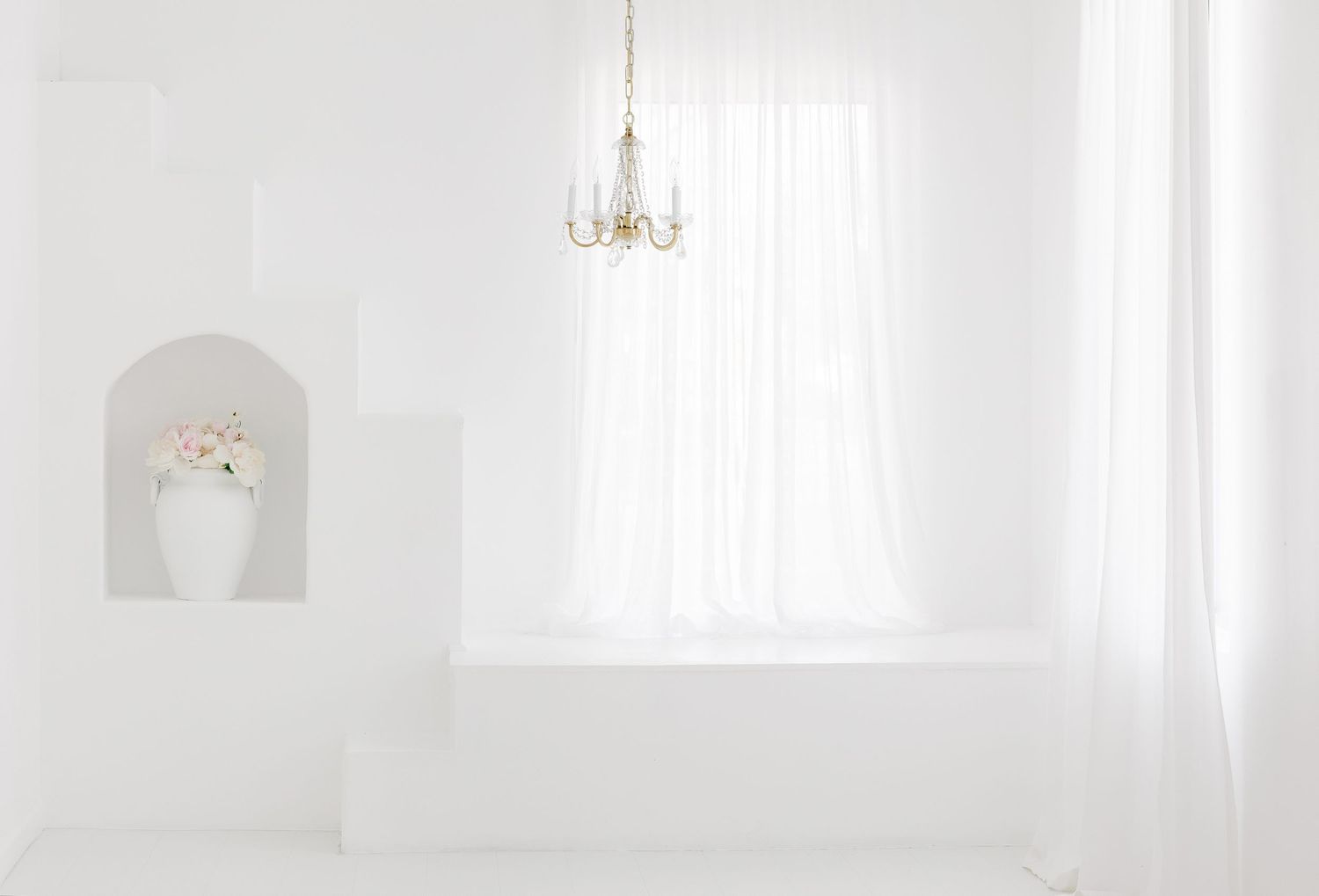 A minimalist white room interior with a small arched alcove and delicate chandelier hanging near a bright window.