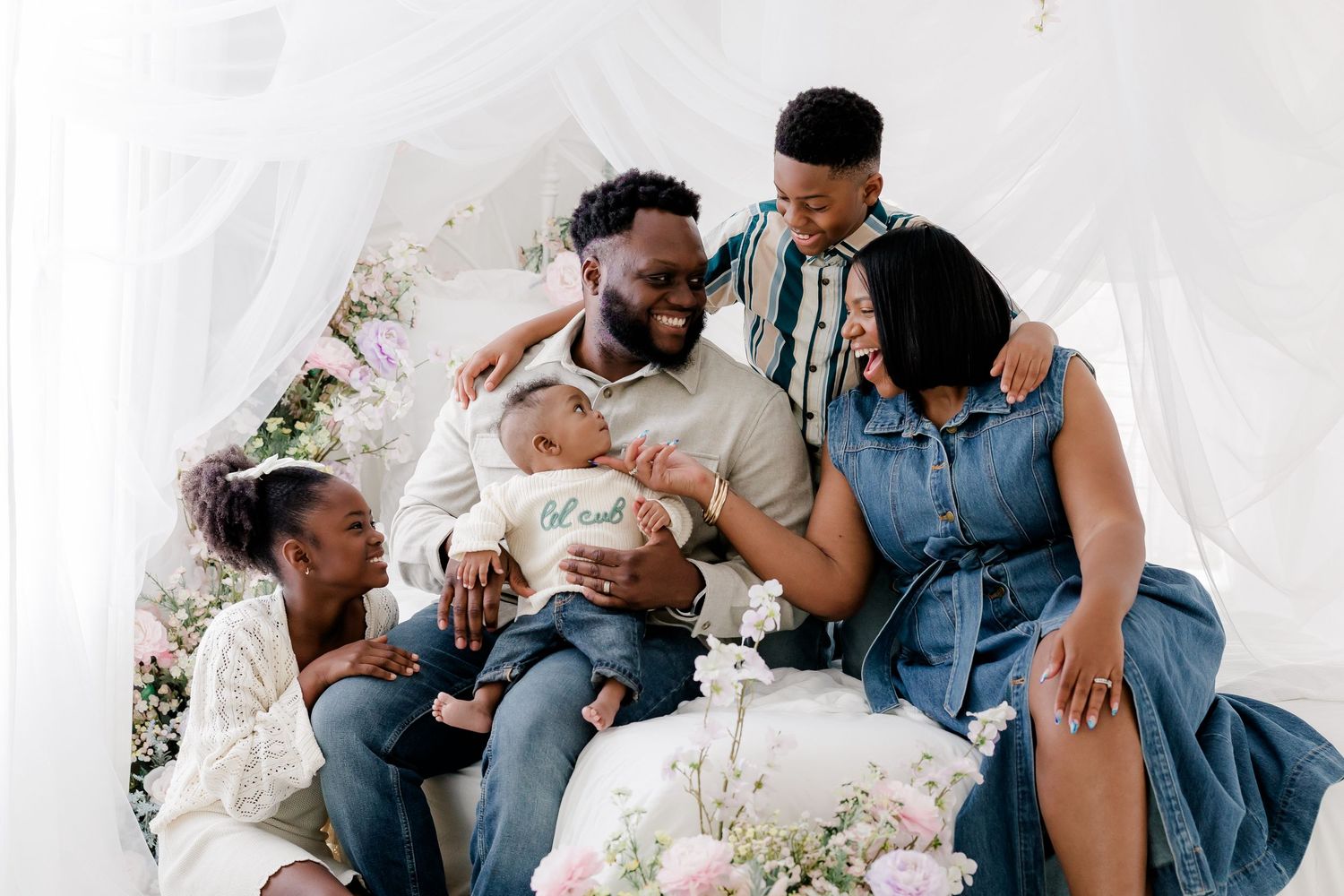 A family sits together on a white couch surrounded by floral arrangements while holding a newborn baby.