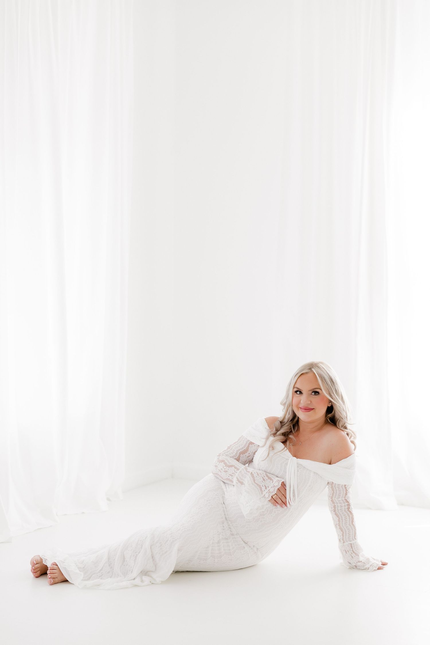 Woman in flowing white dress lying gracefully on white floor in bright minimalist studio setting.