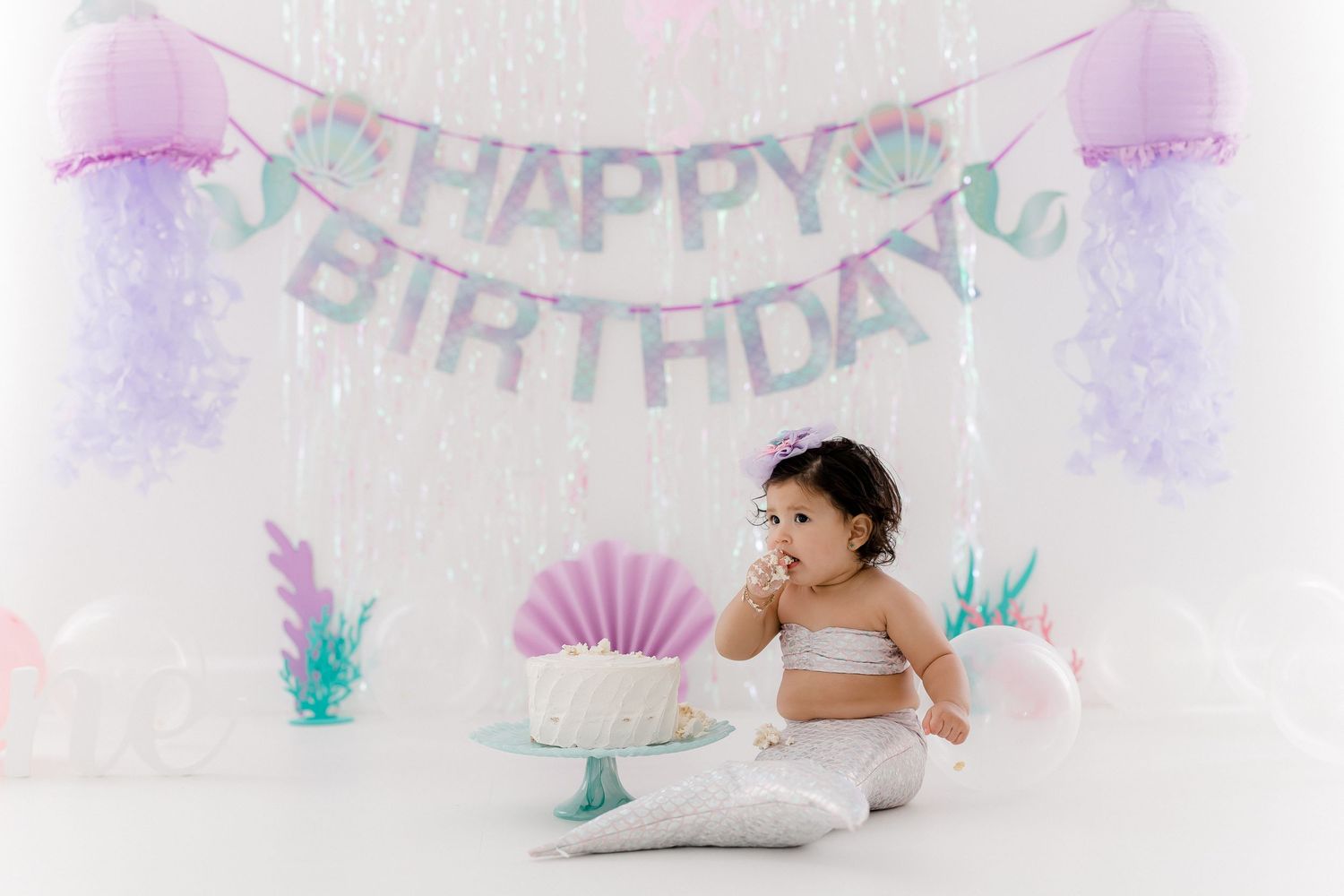 A person in white sits on a floor with pink and teal birthday party decorations and balloons in the background.