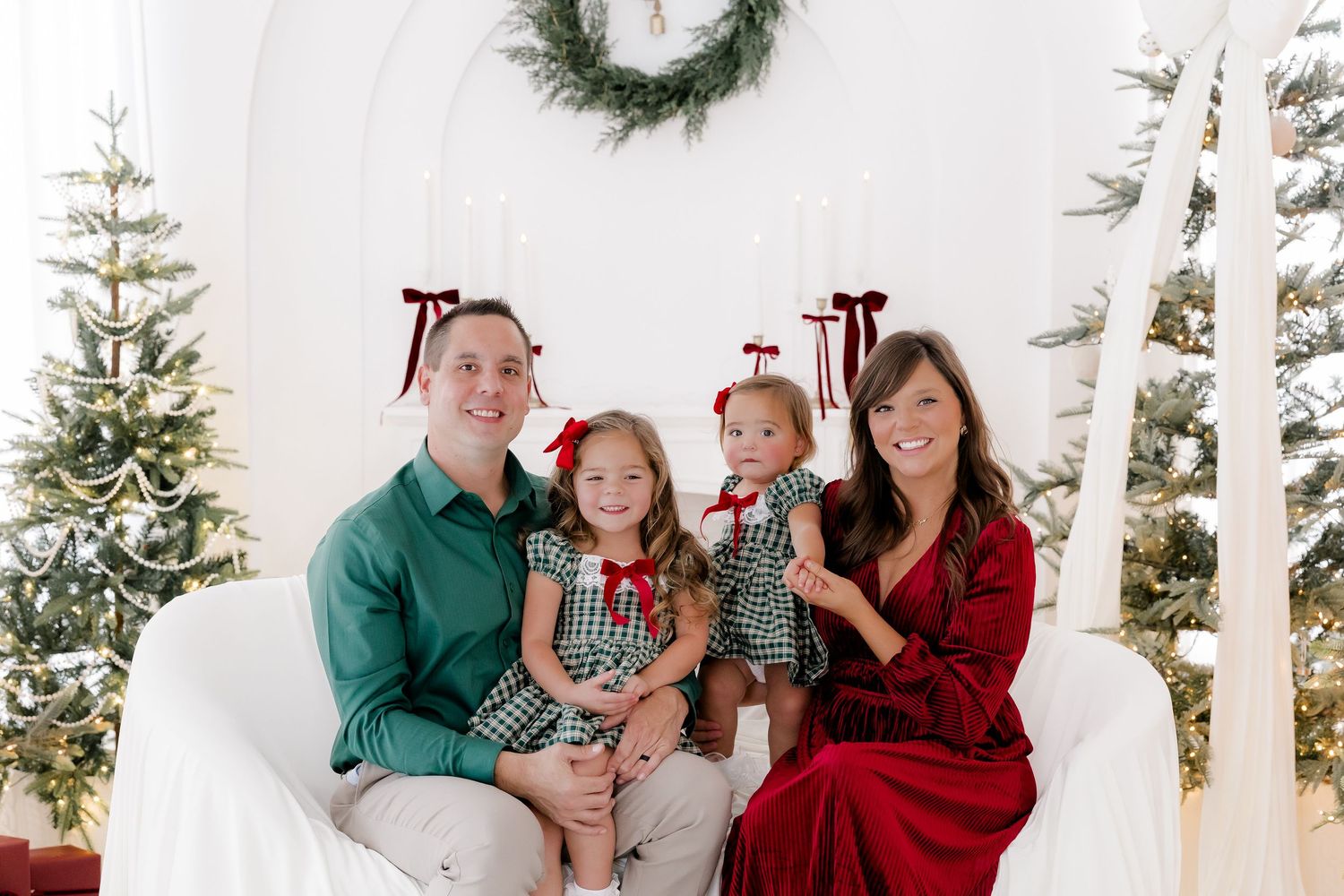 Family of four in festive Christmas attire posing together on white chair with holiday decor and trees.