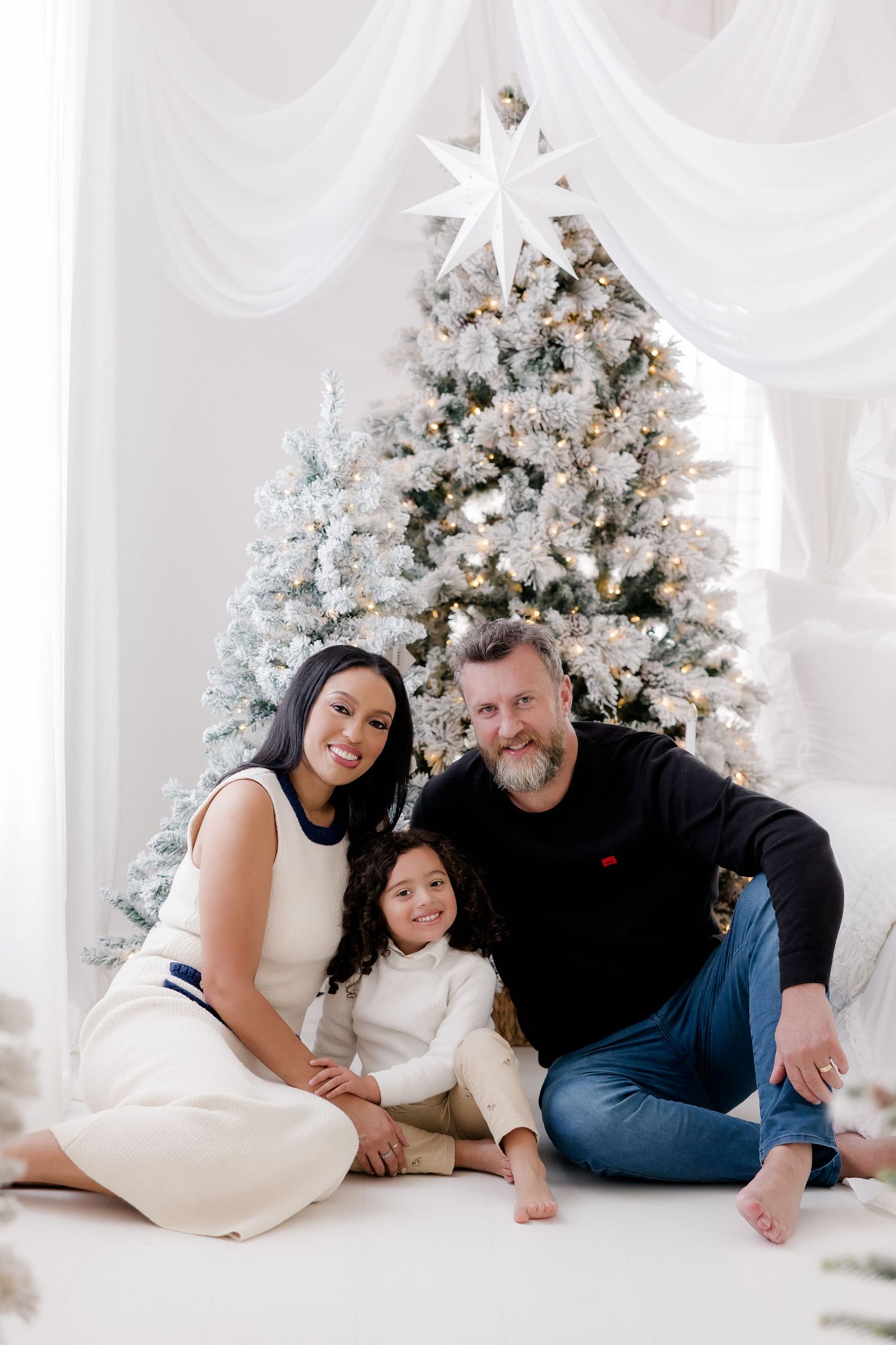Interracial family of three smiling together in front of a white flocked Christmas tree with star topper.