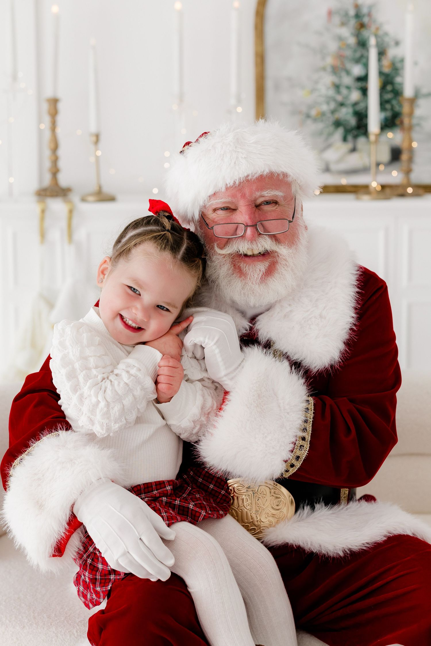A joyful toddler girl sits on Santa Claus's lap, smiling in a festive holiday photo setting.