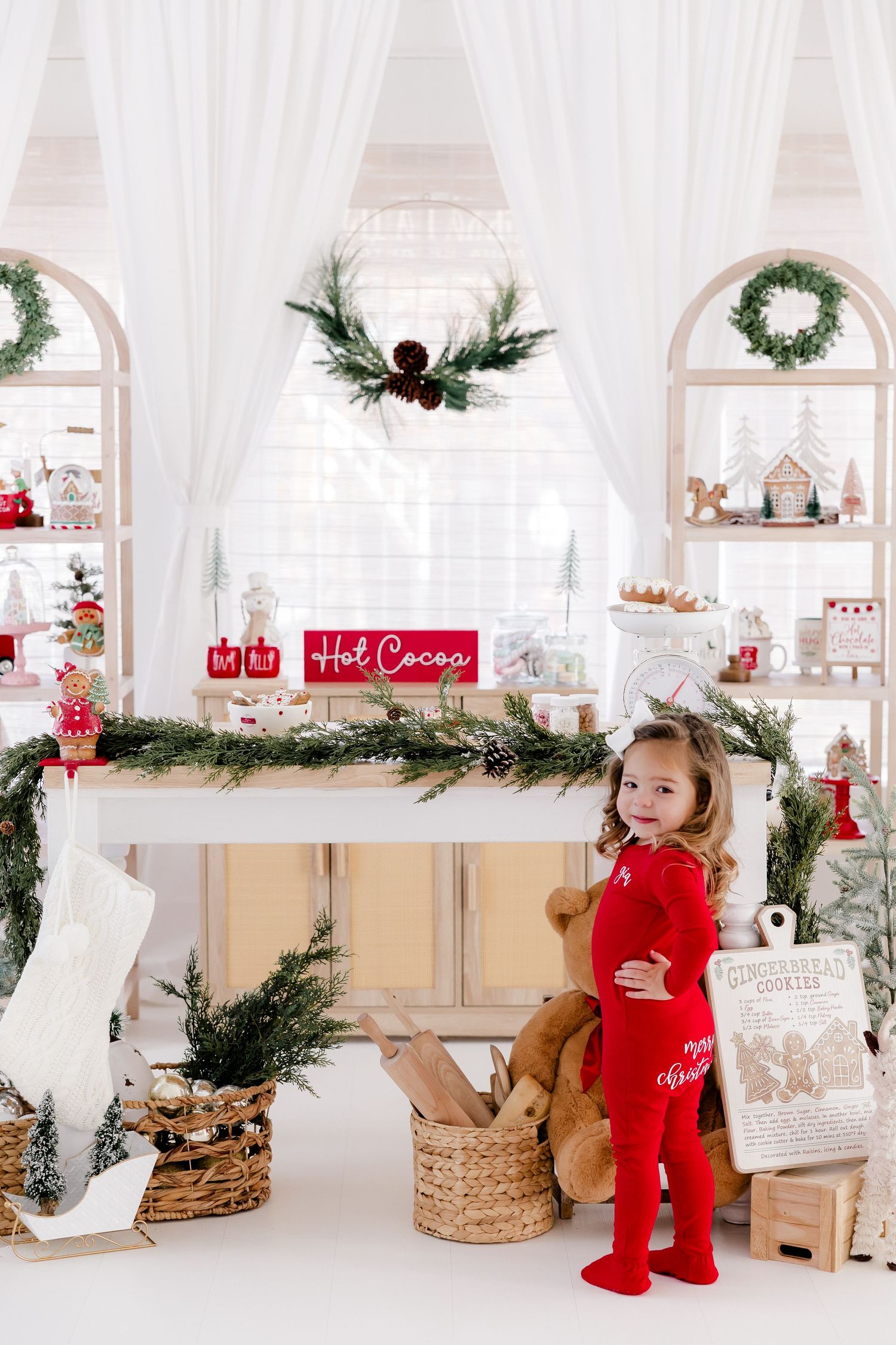 Child in red outfit explores Christmas-themed set with wreaths and holiday decorations.