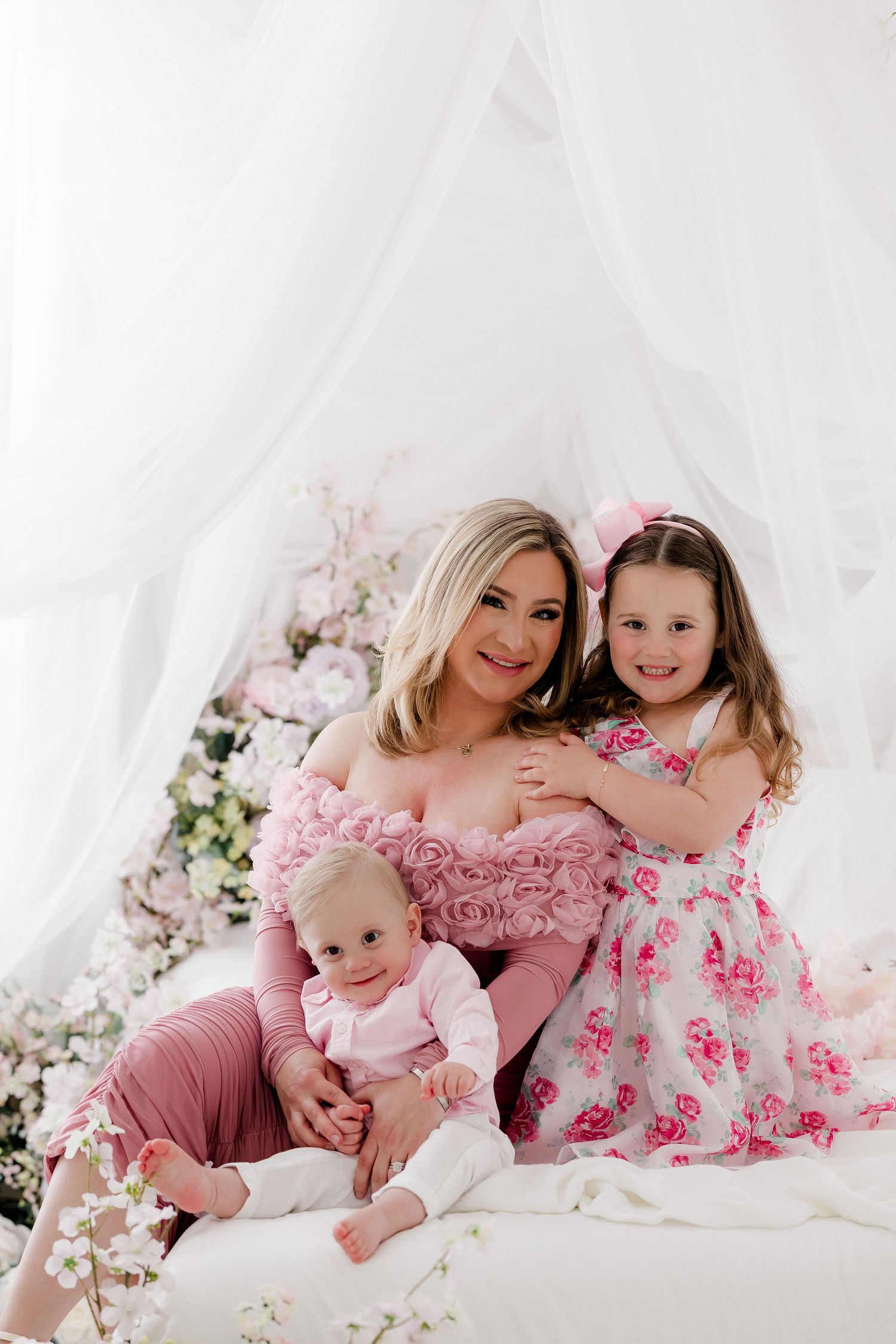 Family portrait in soft pink tones with floral decorations and white draping in elegant studio setting.