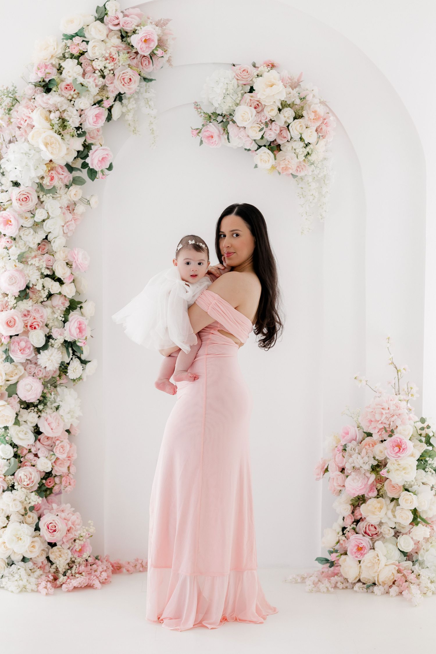 A mother and baby in matching pink dresses pose against a romantic floral backdrop with pink and white roses.