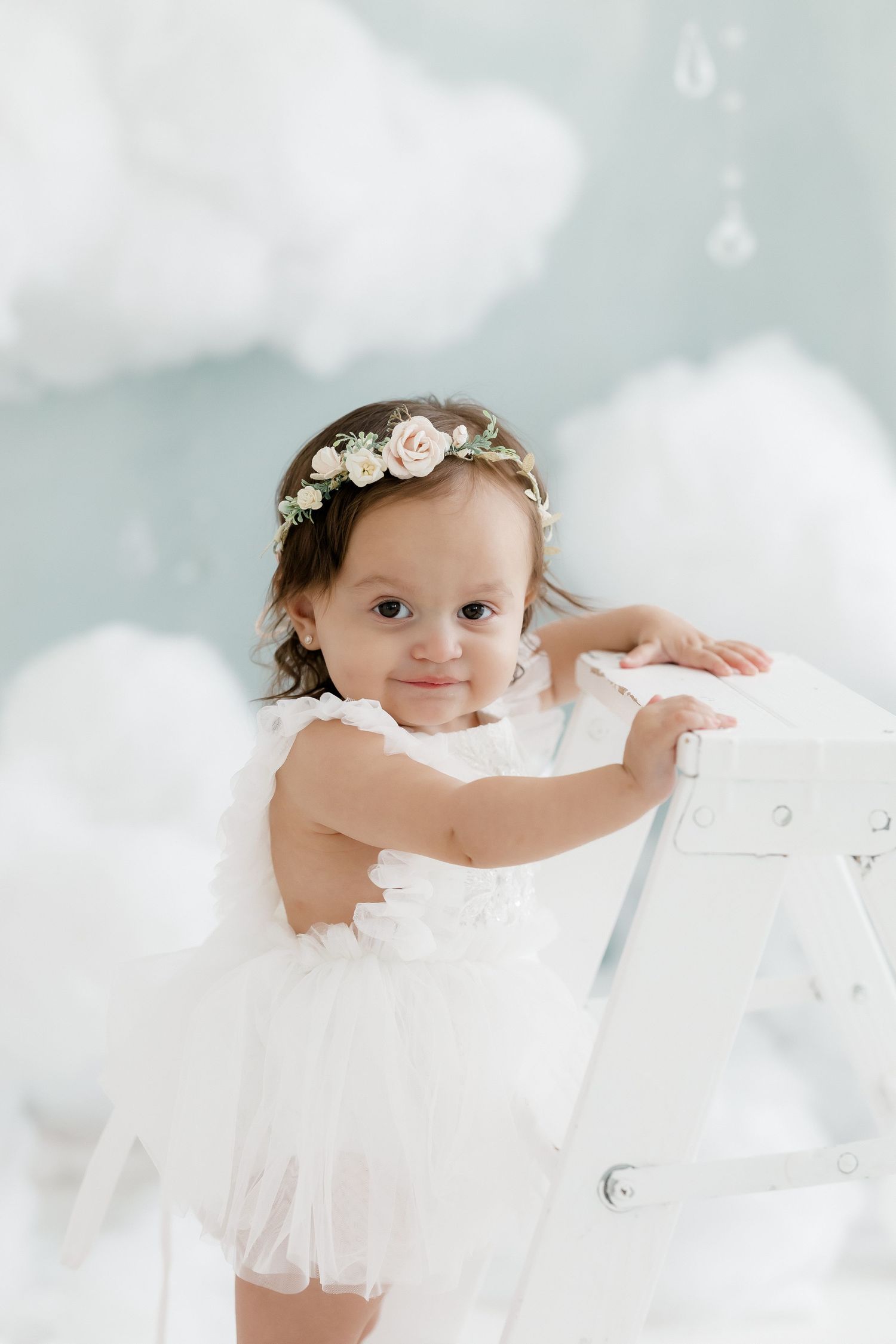 Baby girl in white tutu dress and floral crown smiles while leaning on white ladder with cloud backdrop.