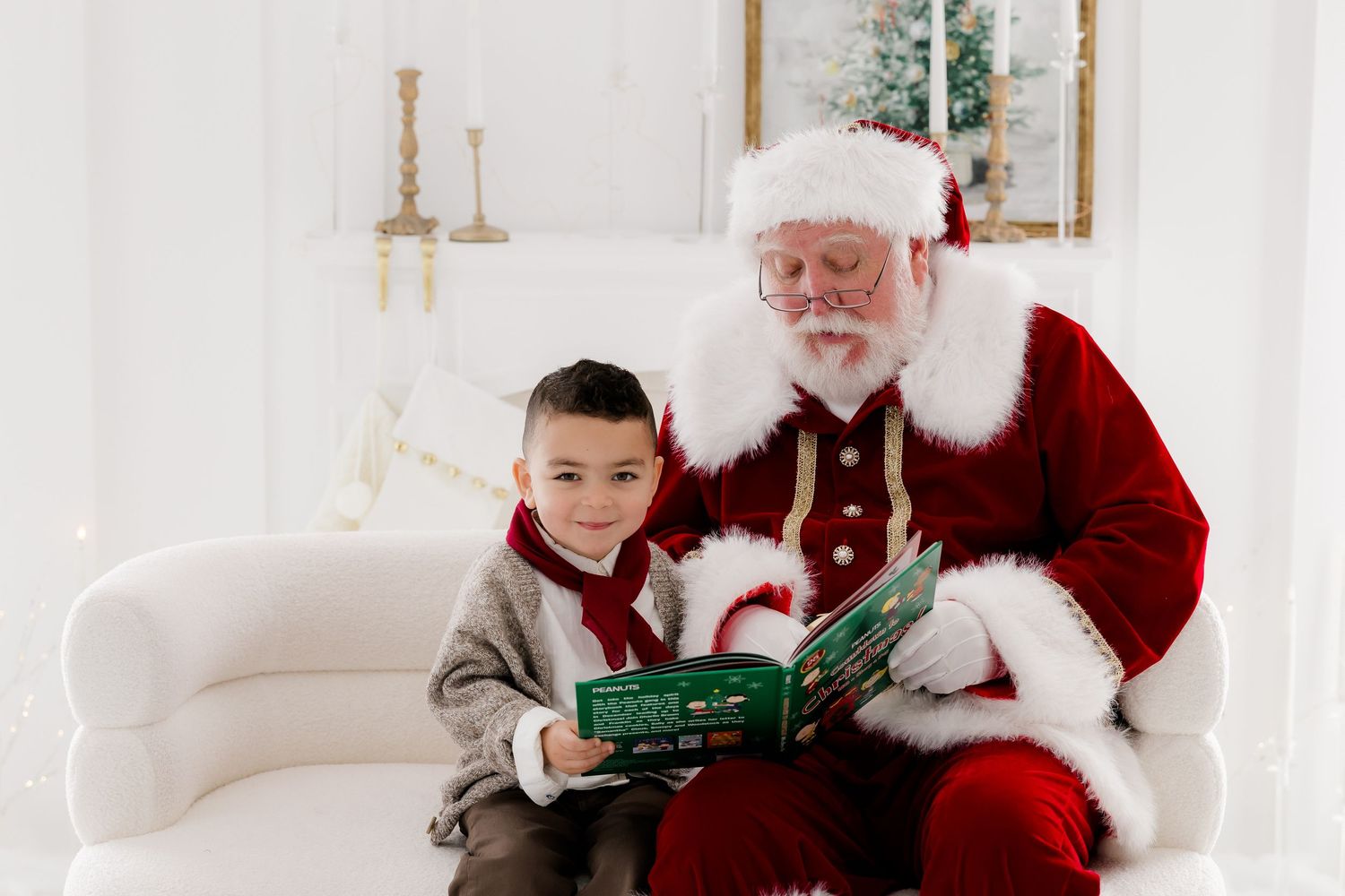 A young boy sits with Santa Claus on a white sofa reading a book in a bright holiday photo studio setting.
