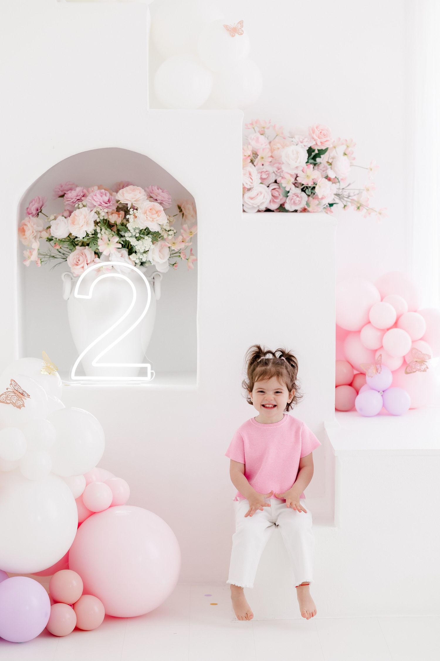 Pink and white birthday party setup with balloons, floral decorations, and a white backdrop in a bright studio setting.